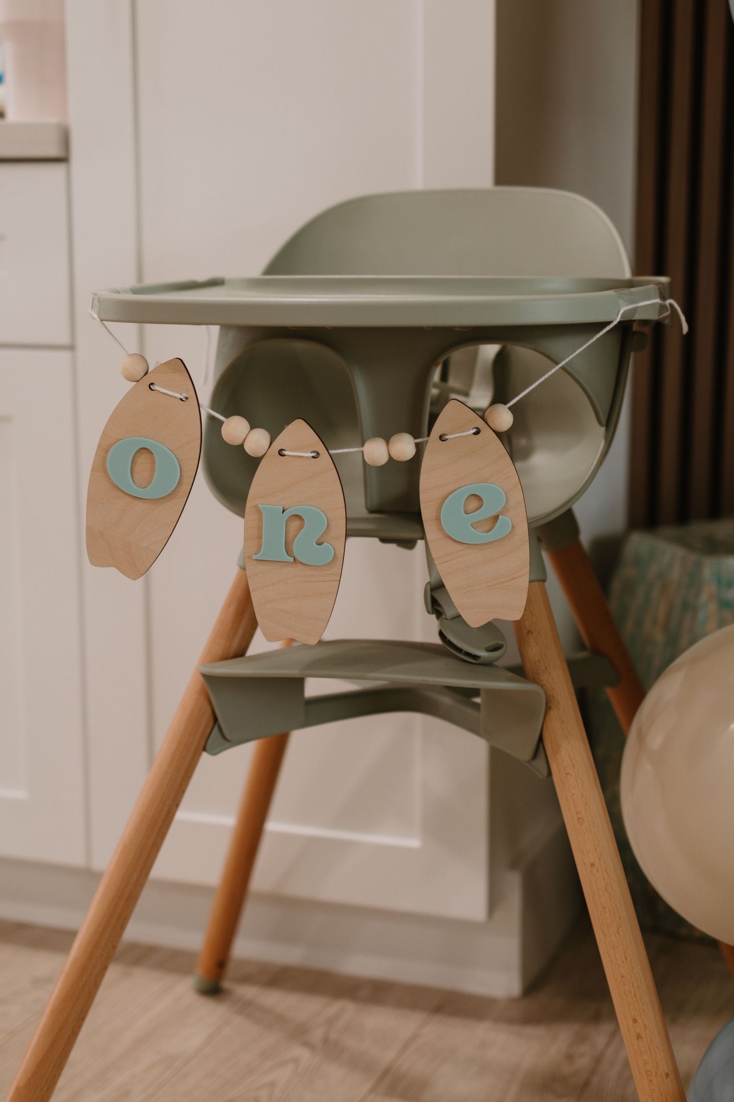 Sage green high chair decorated with wooden surfboard-shaped ONE banner for first birthday.