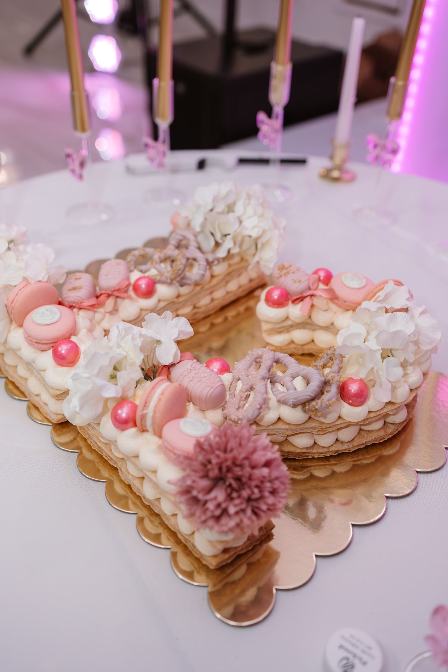 Letter B shaped cream cake decorated with macarons, flowers, and pink pearls on gold board.
