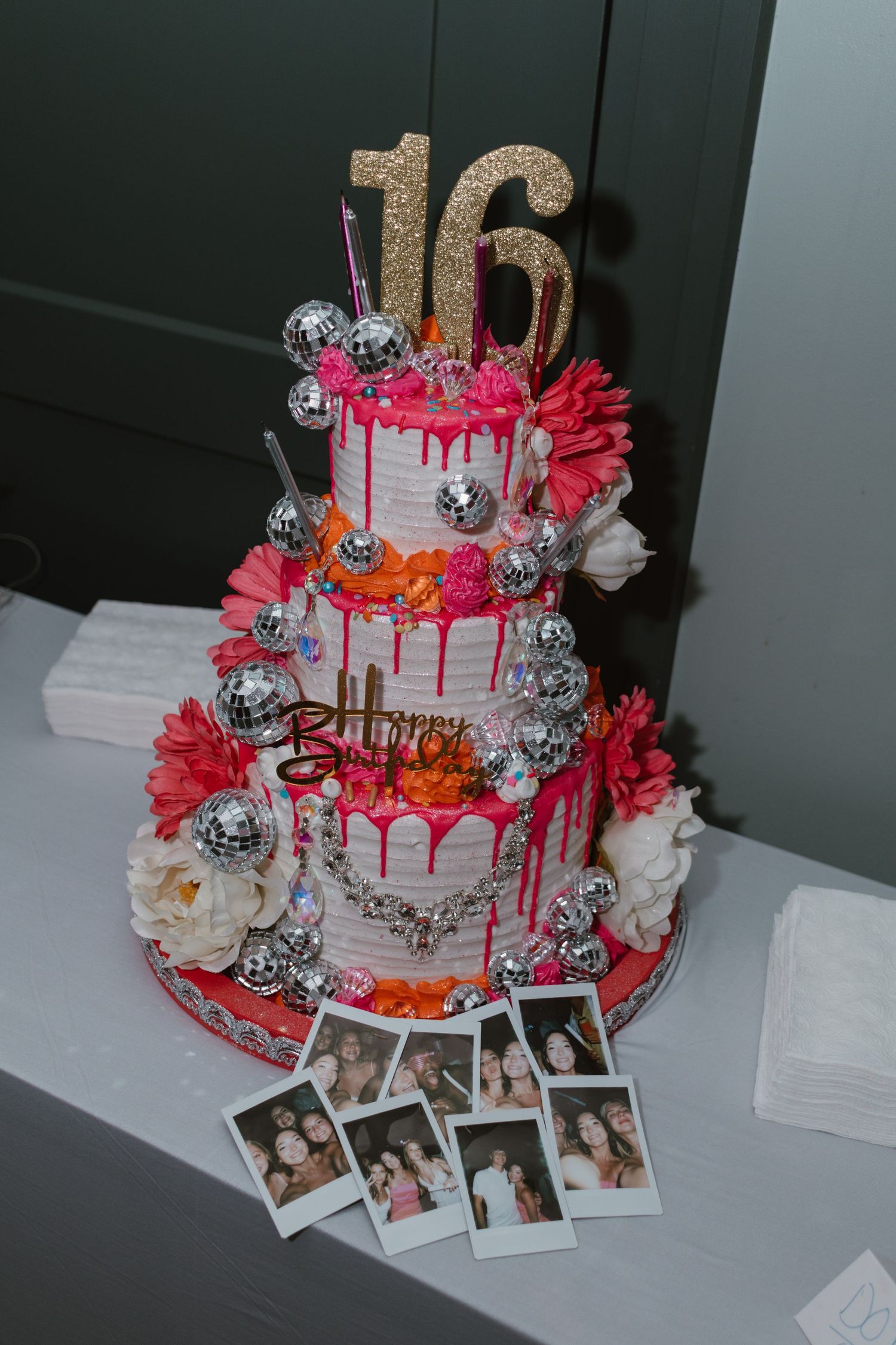 Three-tier Sweet 16 birthday cake with disco balls, pink flowers, gold number 16 topper, and Polaroid photos.