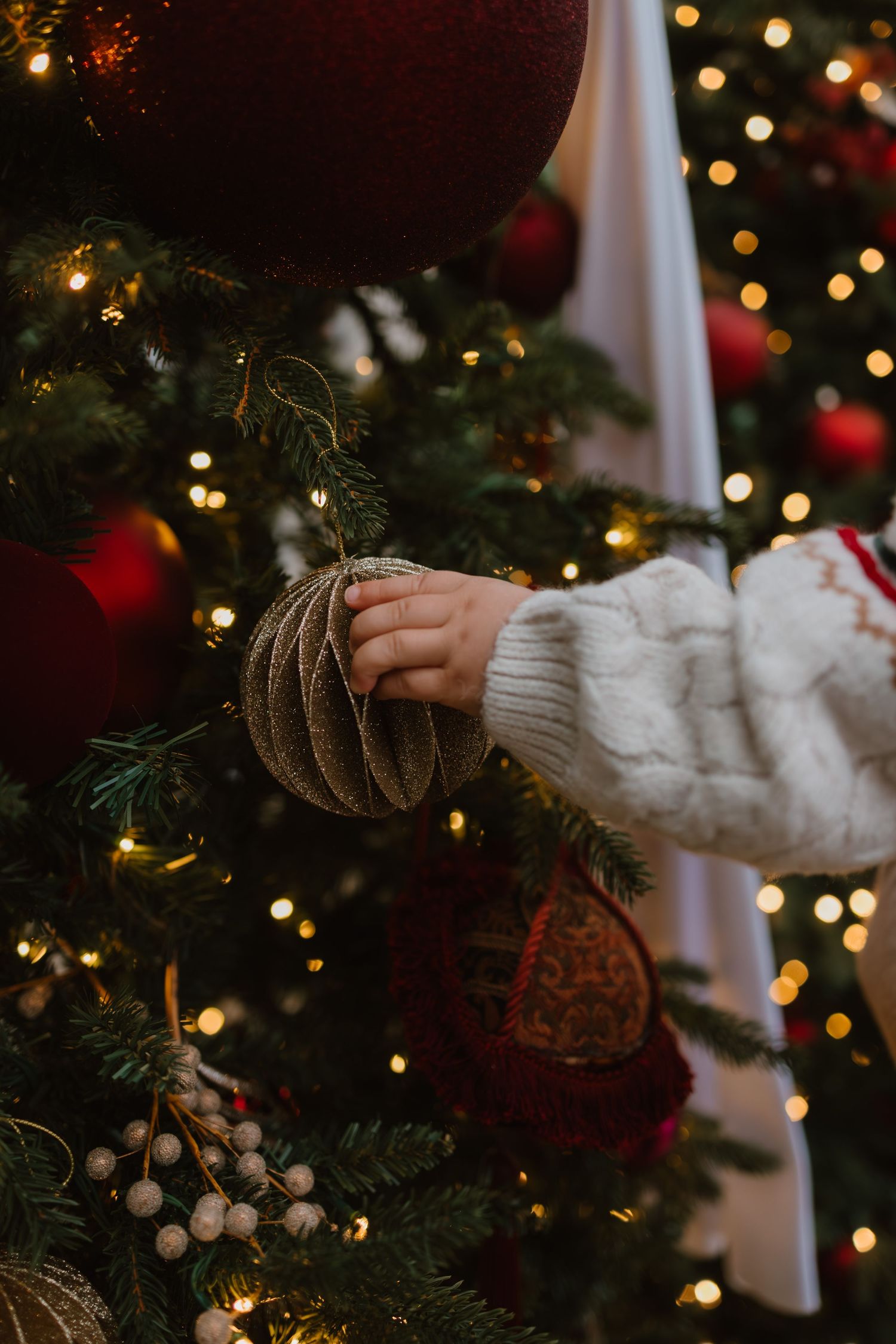 Child's hand in white sweater touching a gold ornament on a decorated Christmas tree with red baubles and warm lights.