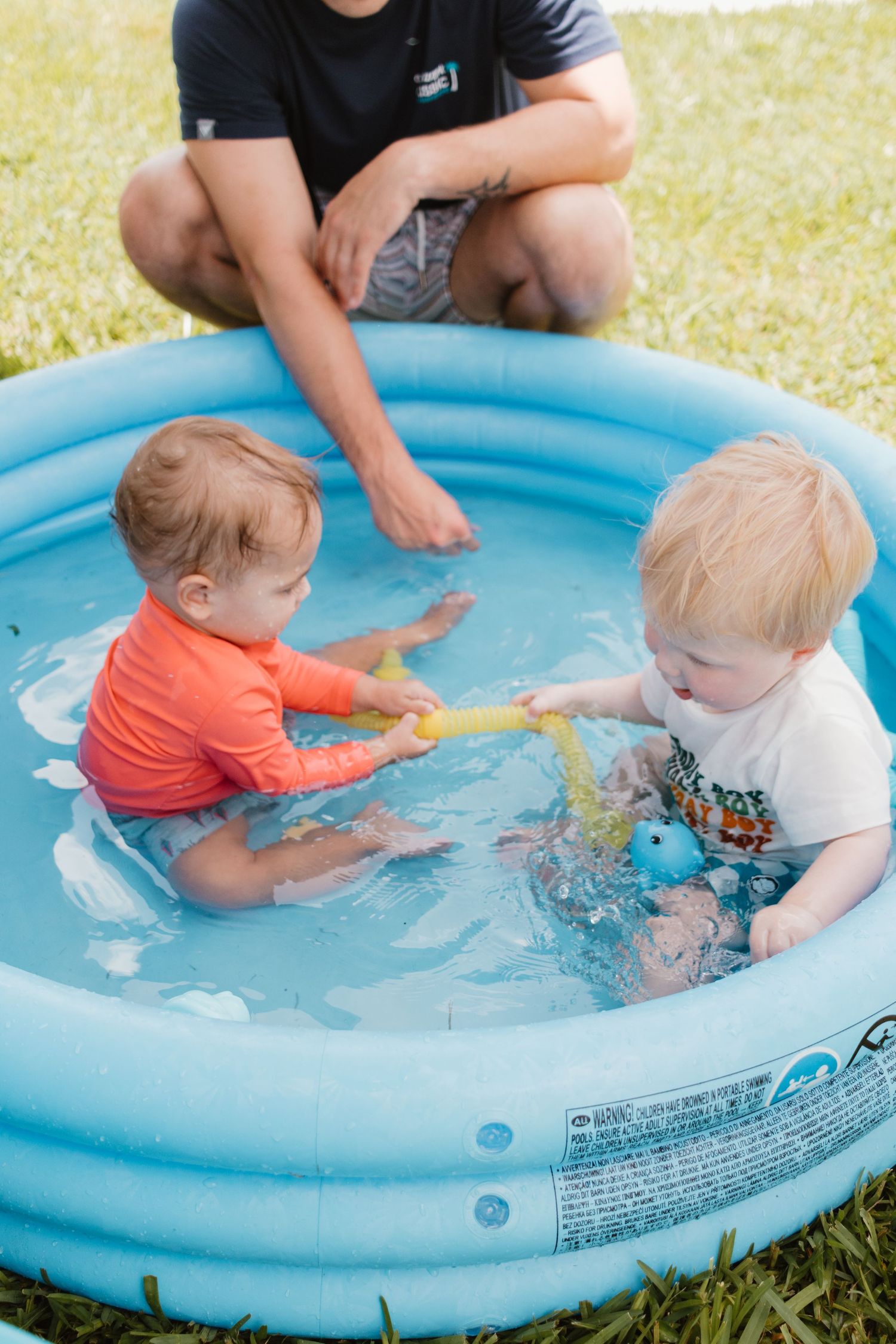 Two babies playing together in blue inflatable kiddie pool supervised by adult at birthday party.