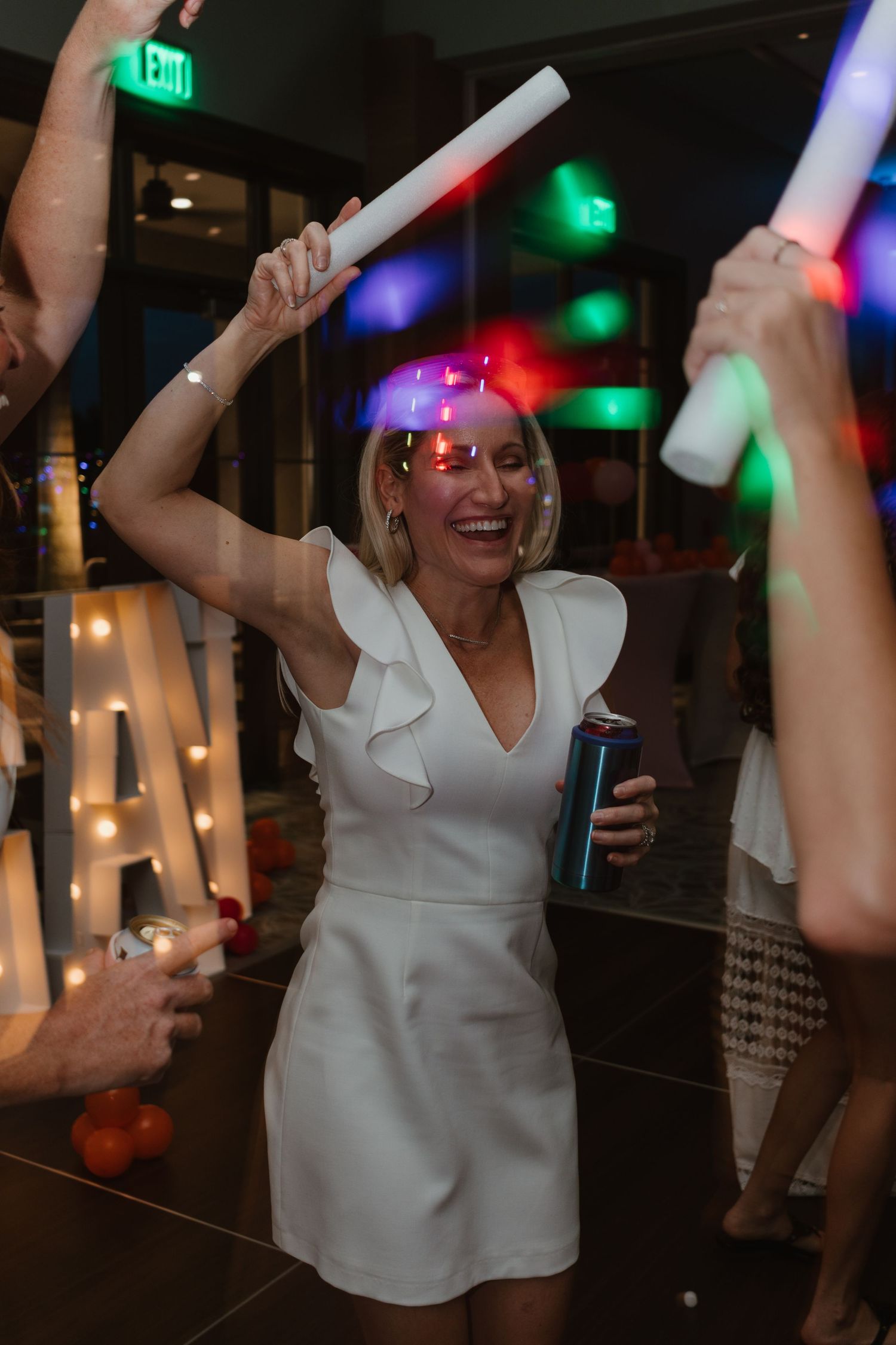 Woman in white dress dancing and smiling at a party holding a can and LED foam stick under colorful lights.
