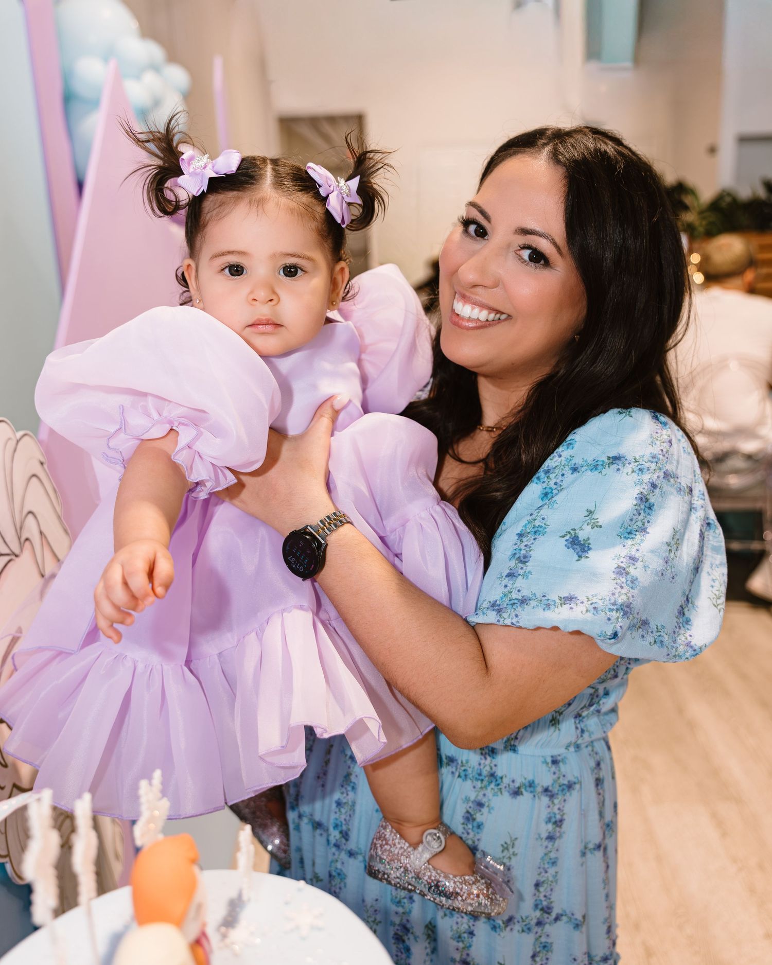 Smiling mother holds baby girl in lavender dress at a Frozen-themed birthday party.