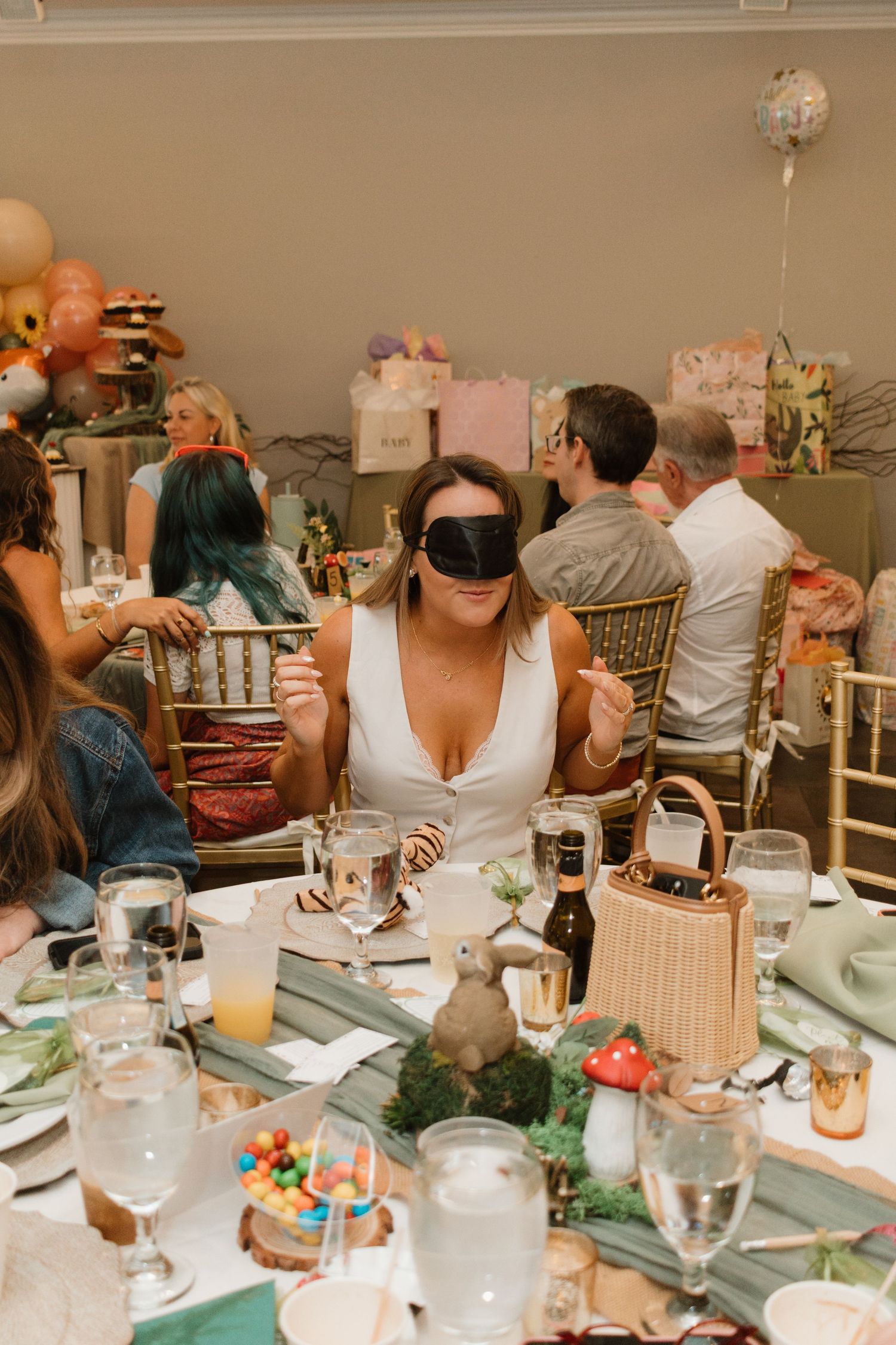 Woman wearing a sleep mask playing a baby shower game at a decorated table with guests around her.
