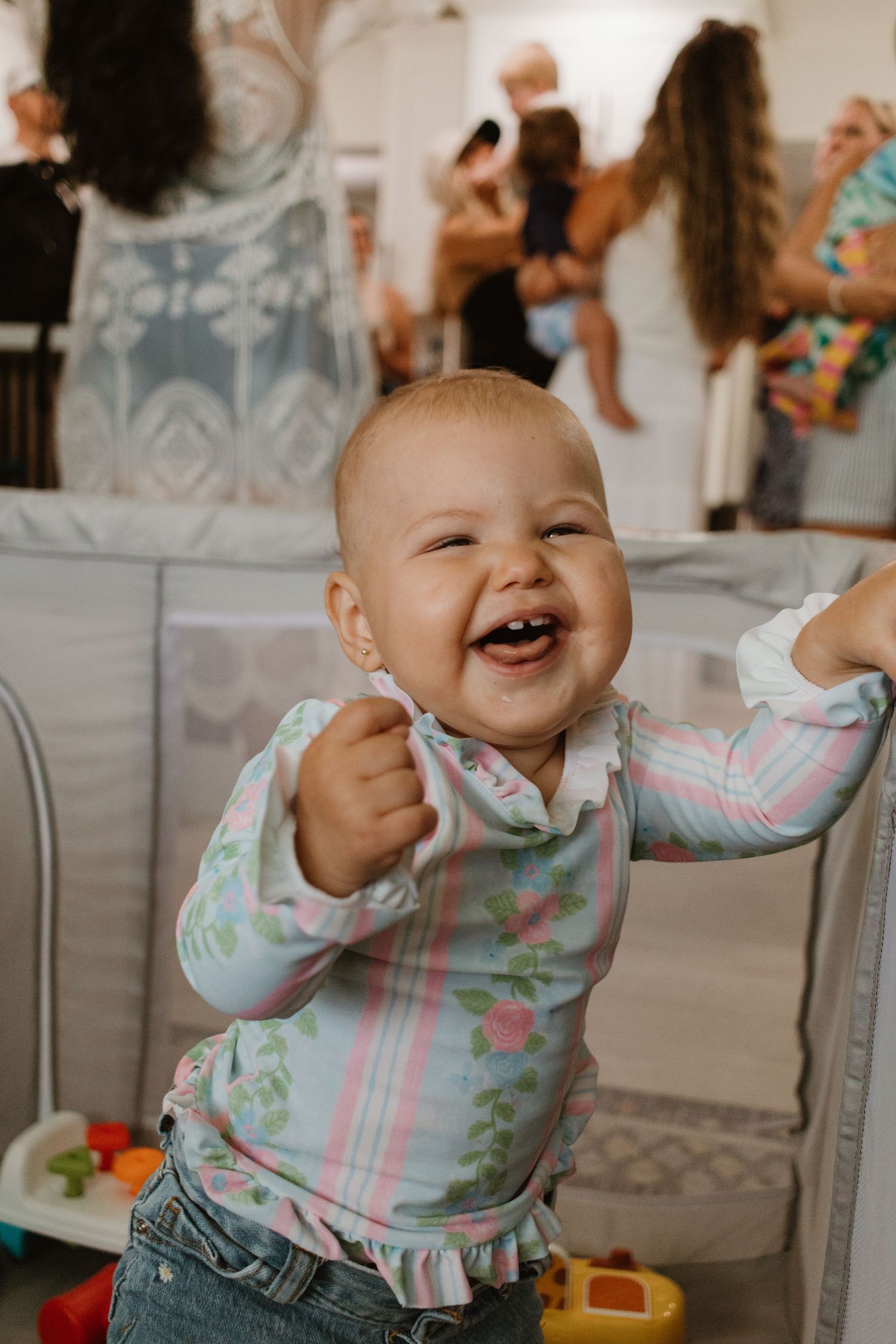 A laughing baby girl in a floral outfit reaches out her arms at an indoor party.
