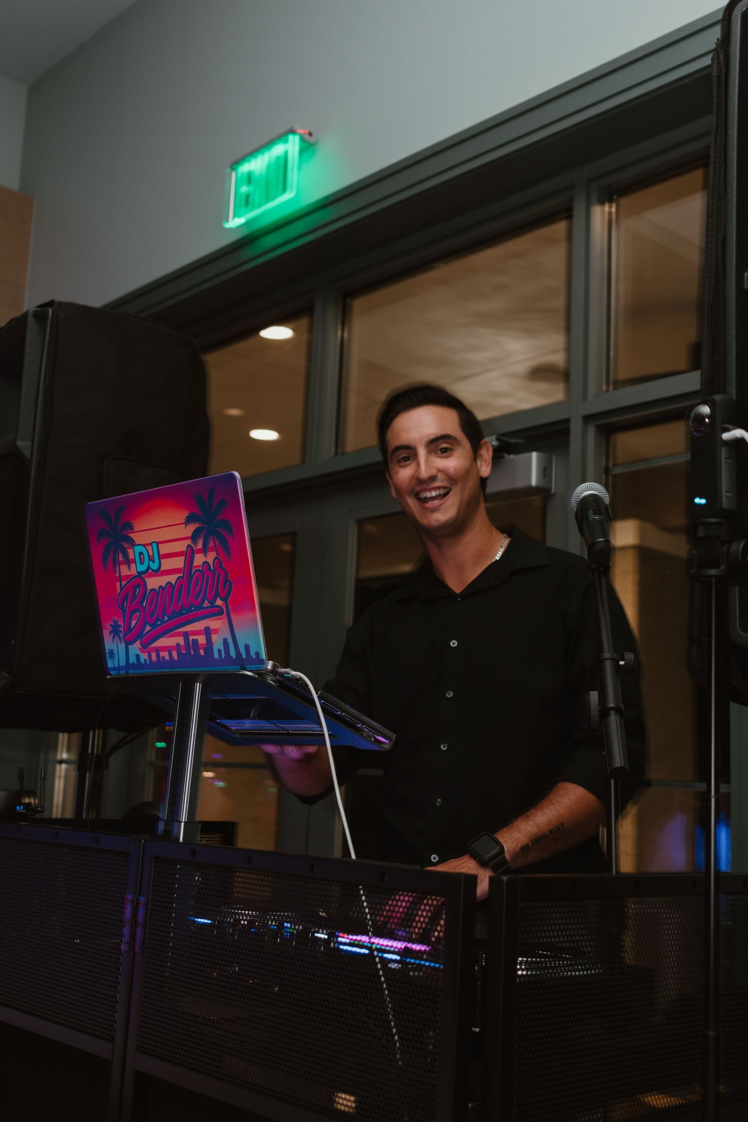 Smiling male DJ standing behind DJ equipment with colorful neon sign at an indoor event.