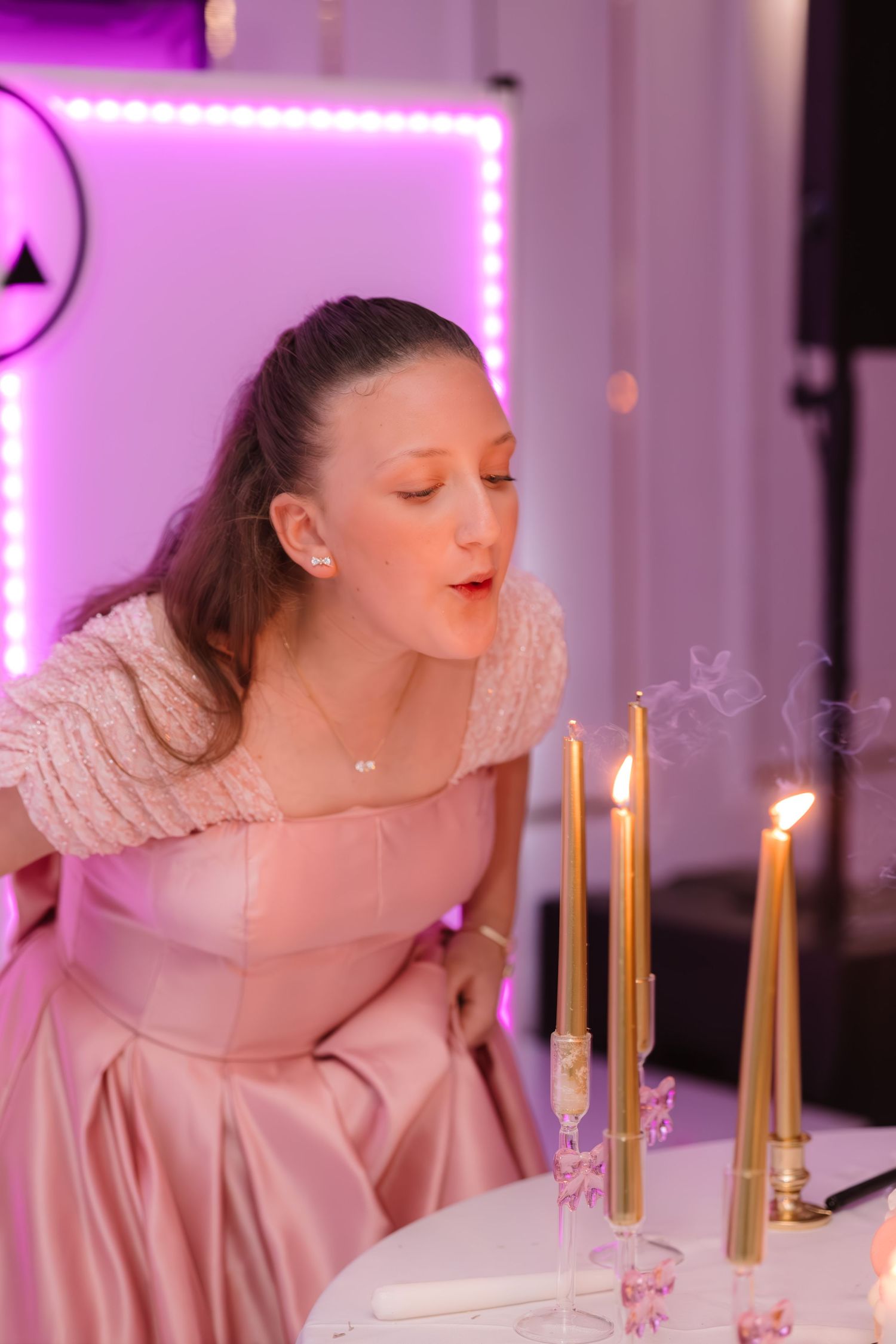 Young woman in pink dress blowing out gold taper candles at a birthday celebration.