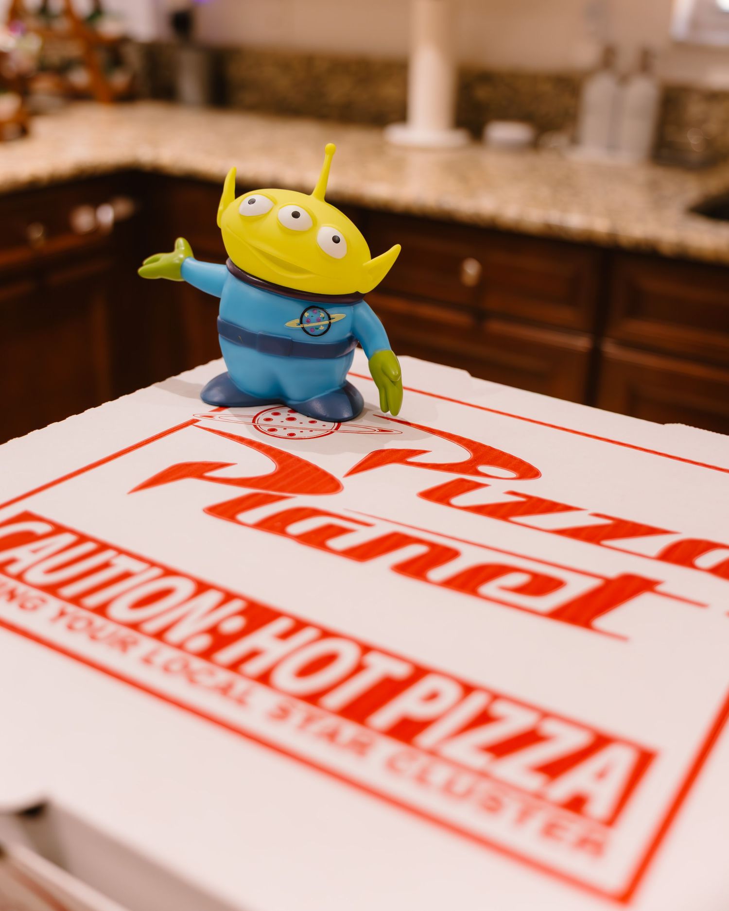 Toy Story alien figurine posed on Pizza Planet themed pizza box at birthday celebration.