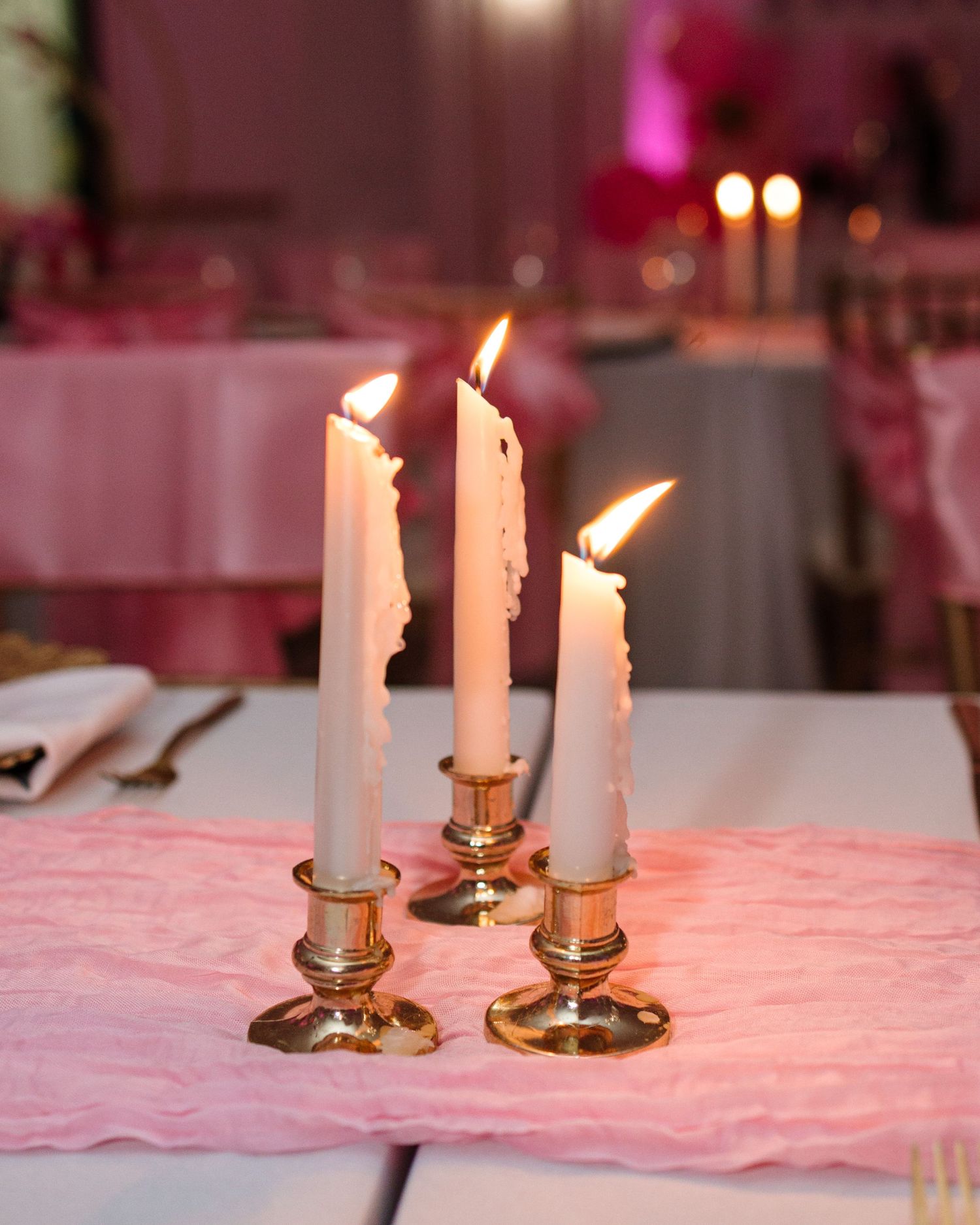 Three white taper candles in gold candlesticks on pink linen tablecloth at elegant party.