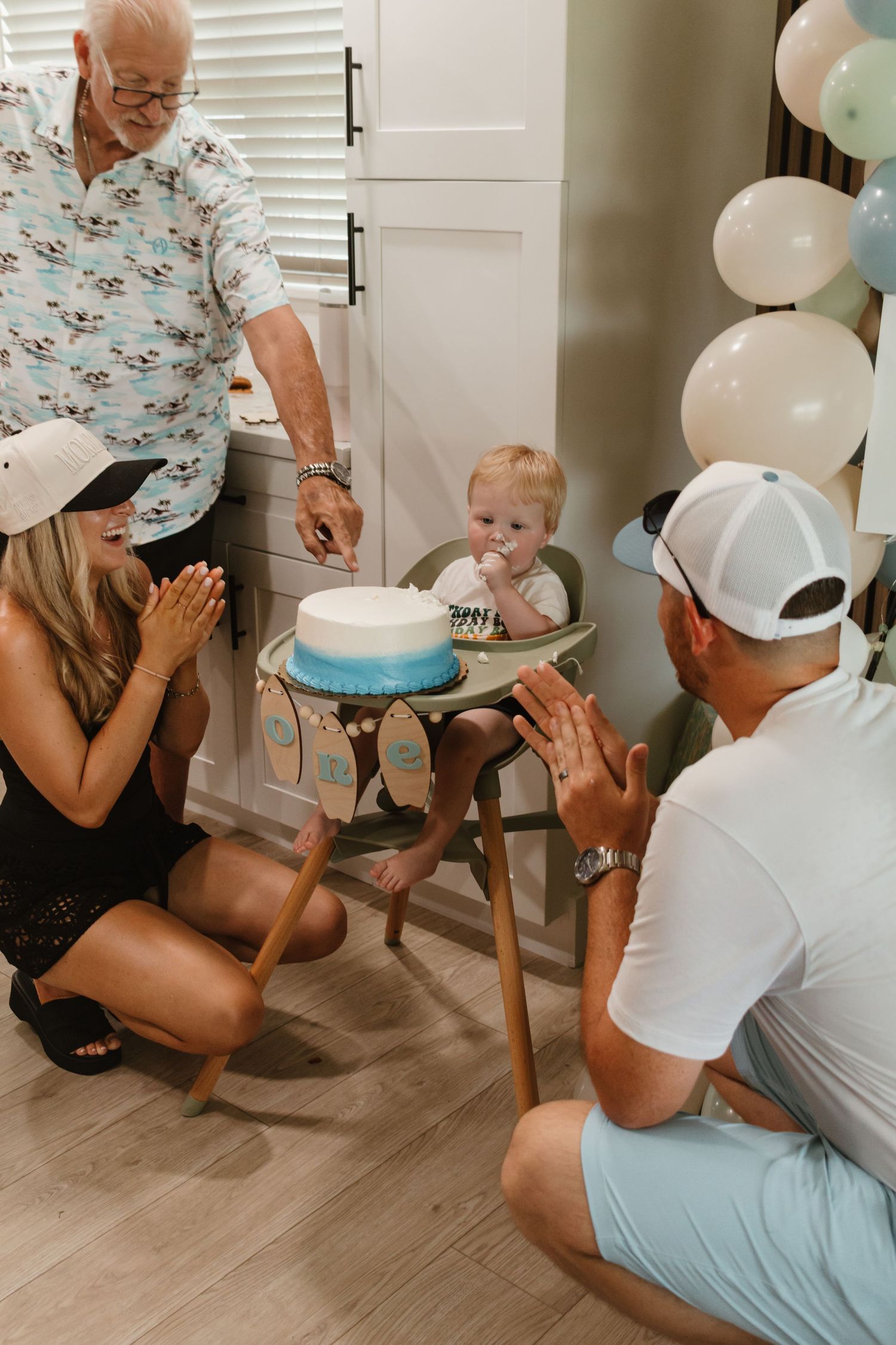 Baby boy enjoying his first birthday cake in high chair surrounded by family and balloons.