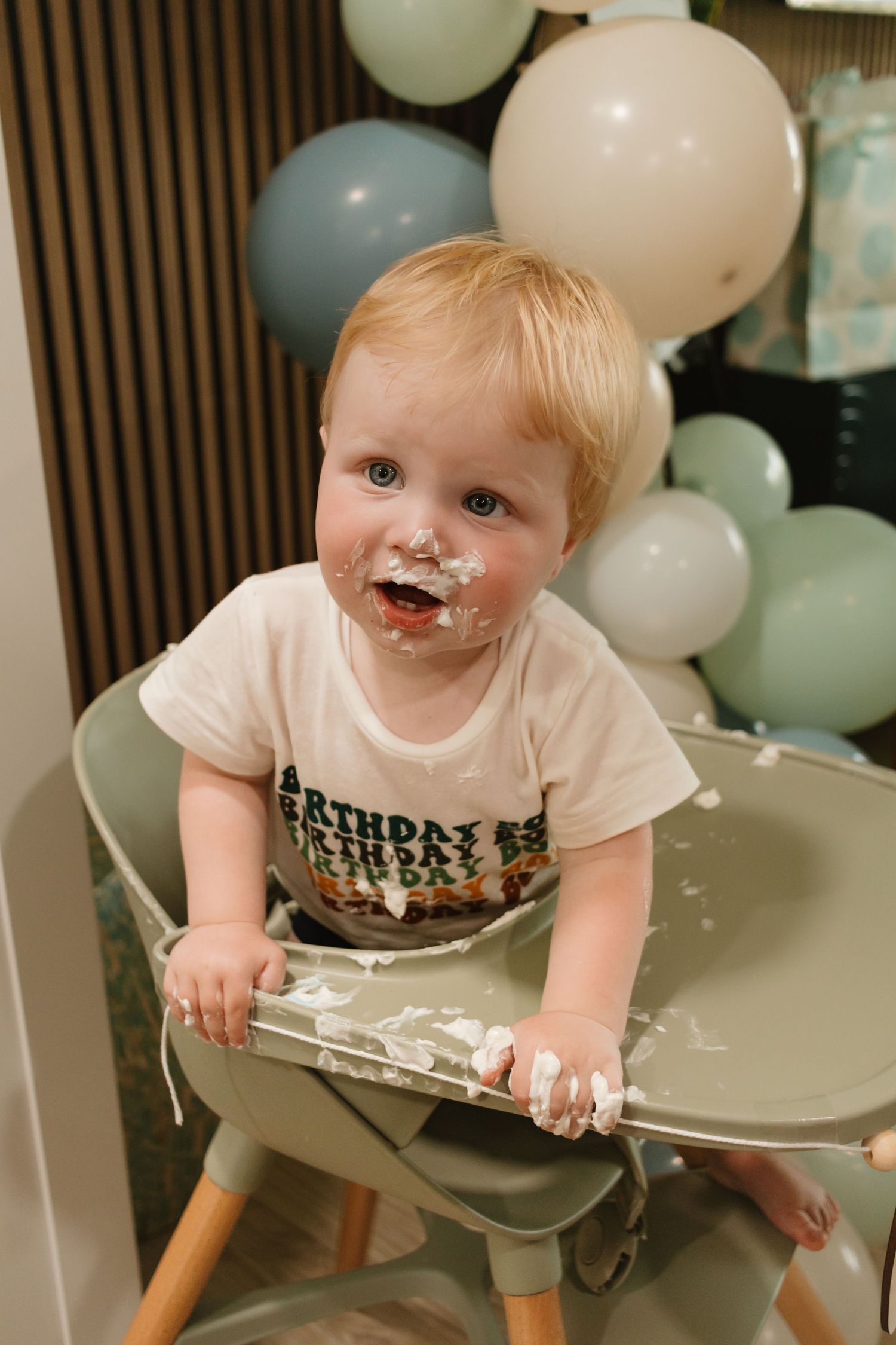 Adorable baby boy covered in white birthday cake frosting sitting in high chair with balloons.