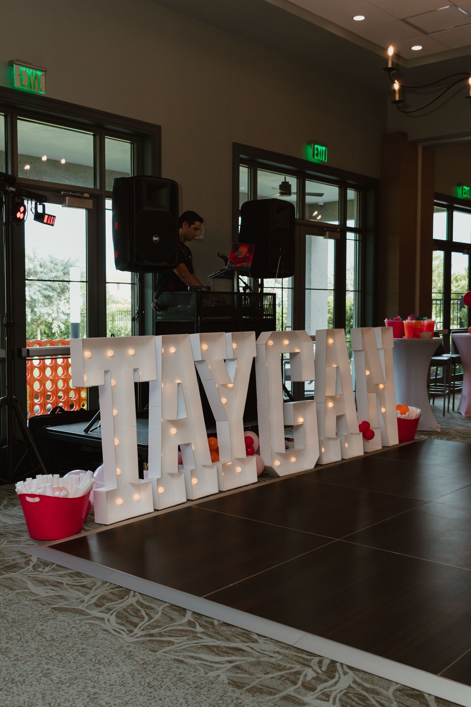 Illuminated marquee letters spelling TAYGAN on a dance floor with a DJ setup and red buckets in the background.