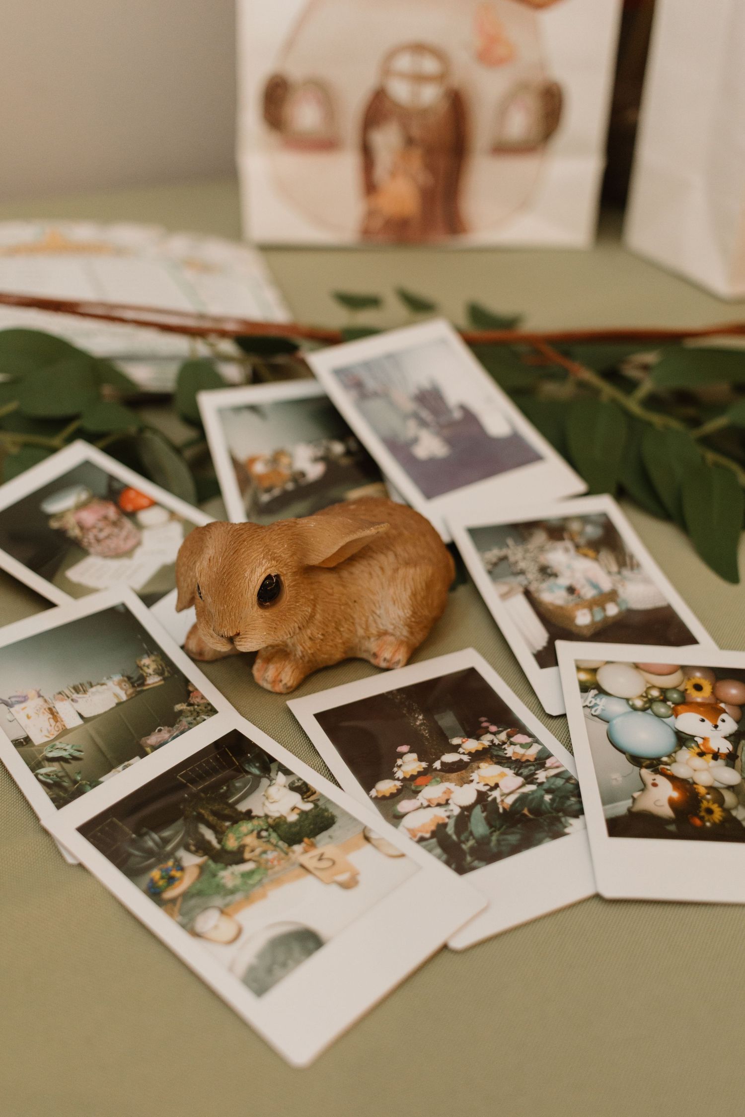 Ceramic bunny figurine surrounded by Polaroid photos and green leaves on a sage green table surface.