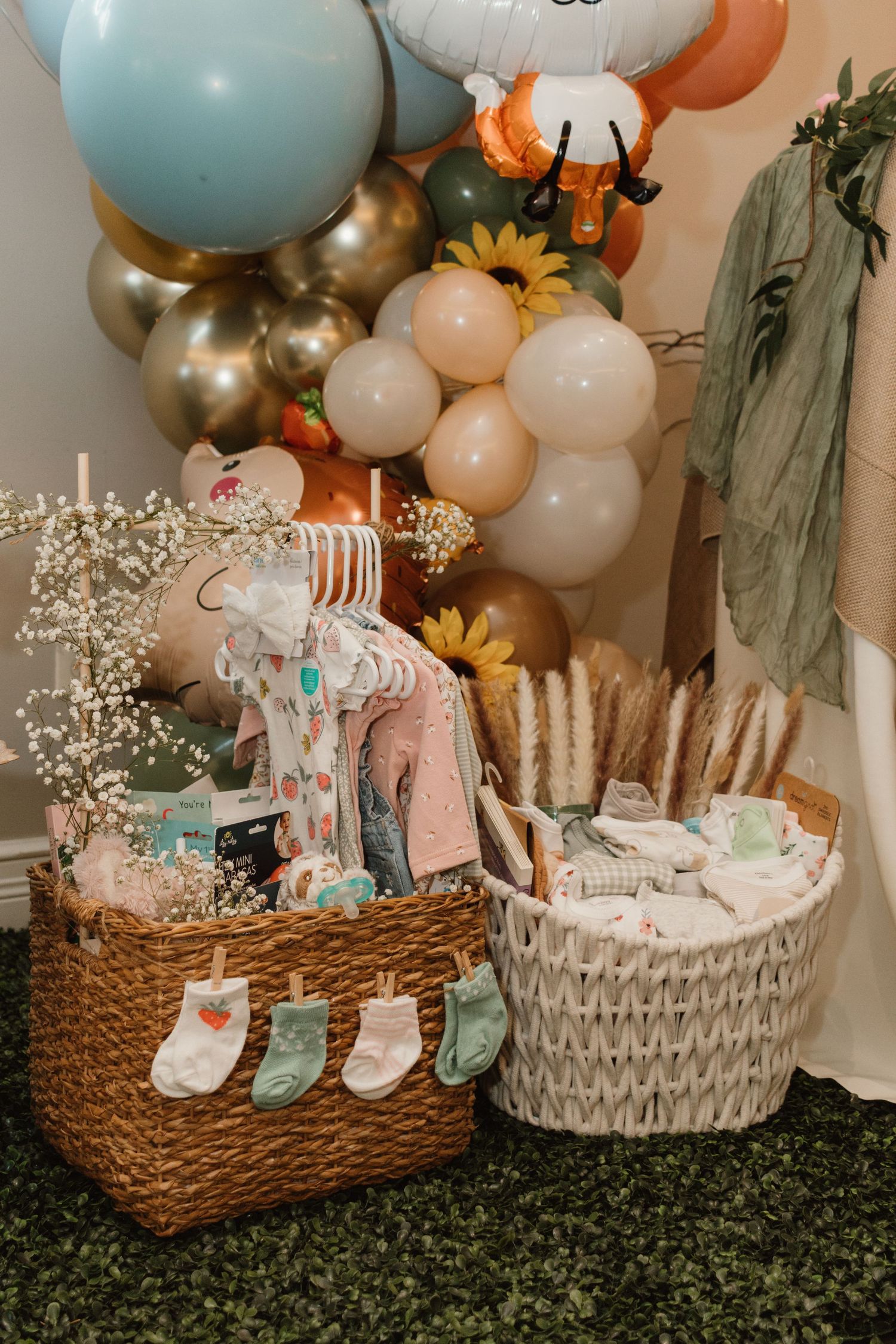 Two wicker baskets filled with baby clothes and essentials displayed beneath a colorful balloon garland with sunflowers.
