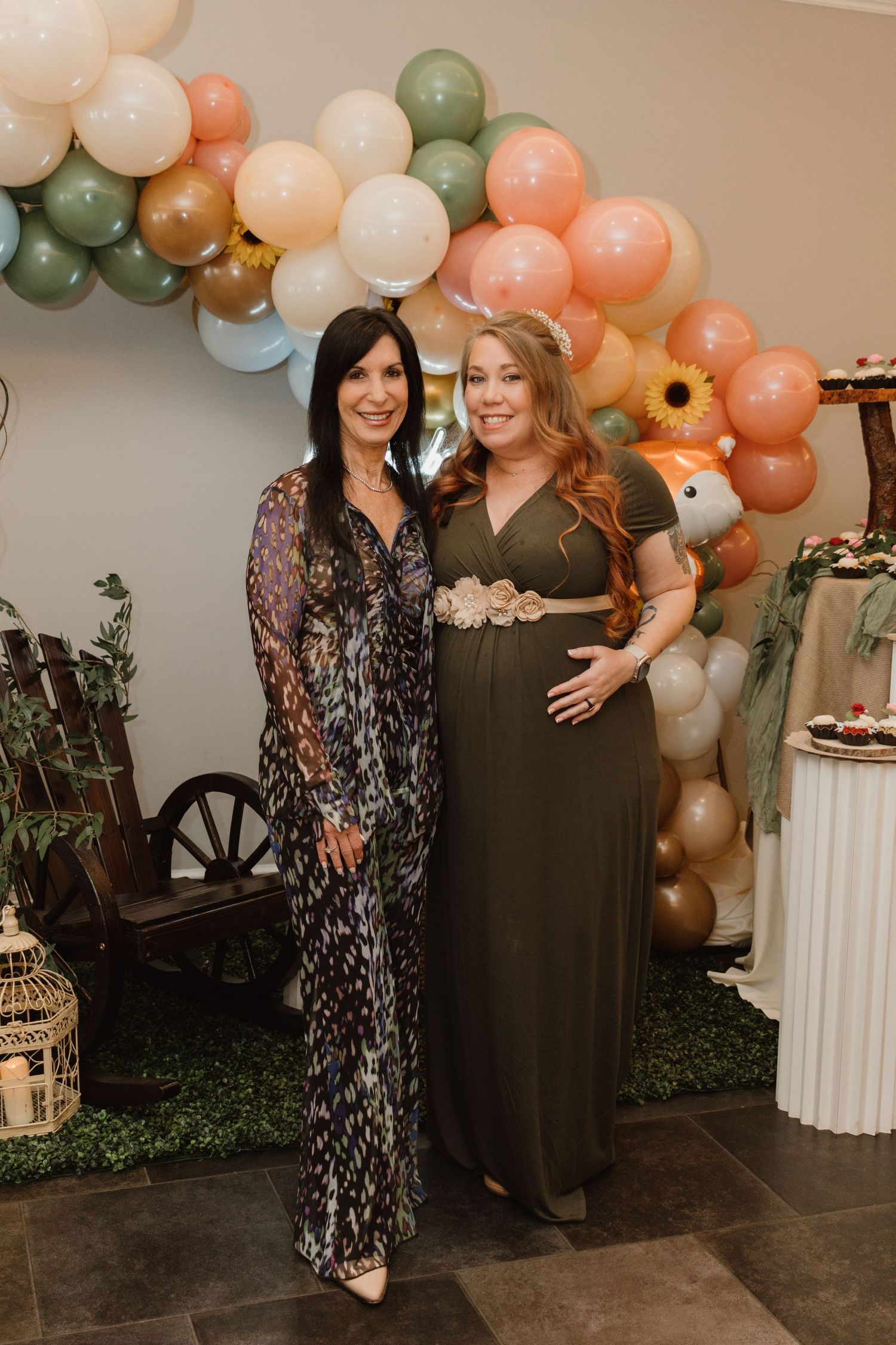 Two women posing together at a woodland-themed baby shower, one in an olive maternity dress holding her bump.