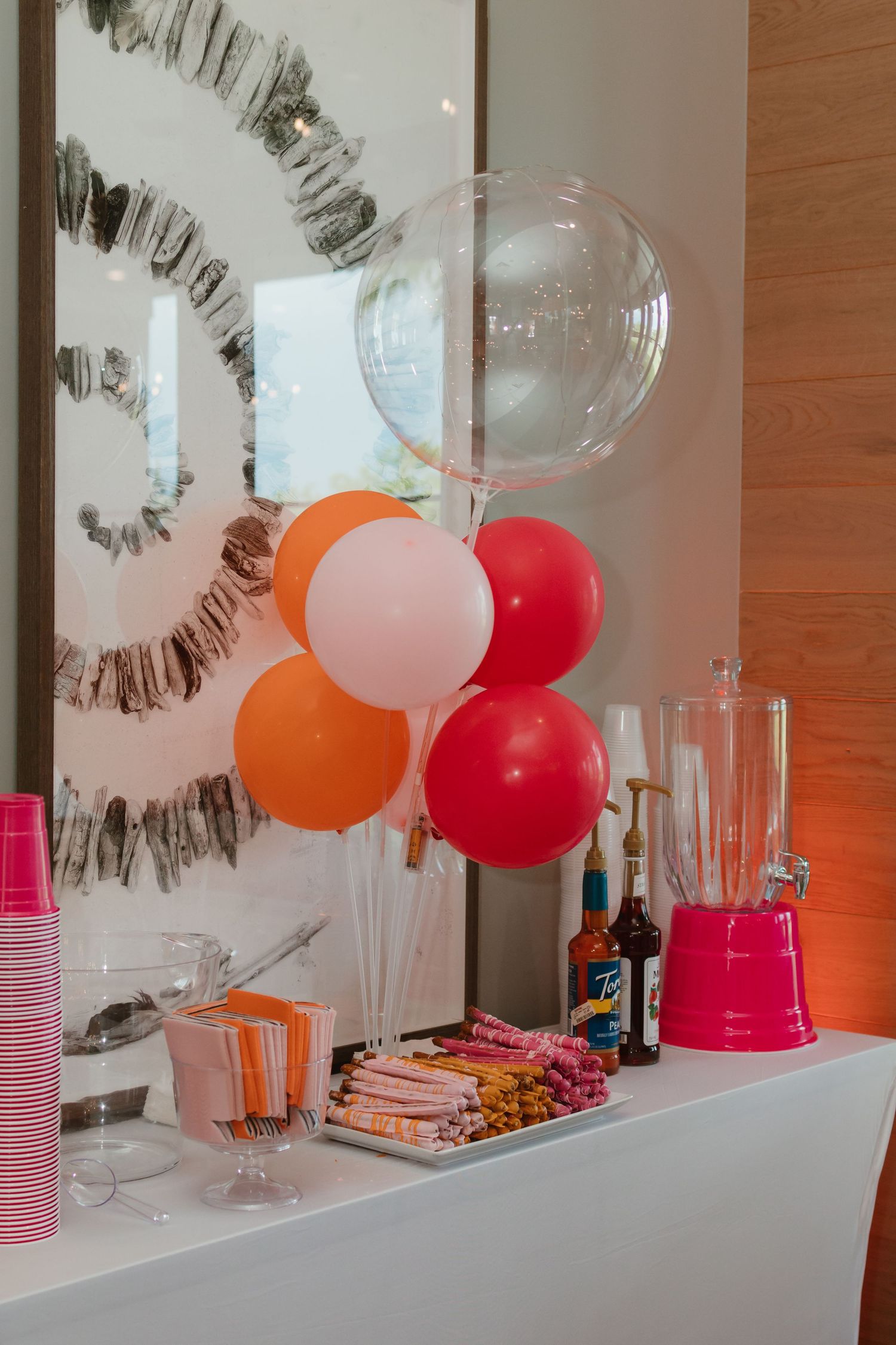 Colorful party drink station with orange, pink, and red balloons, cups, straws, and beverage bottles on a white counter.