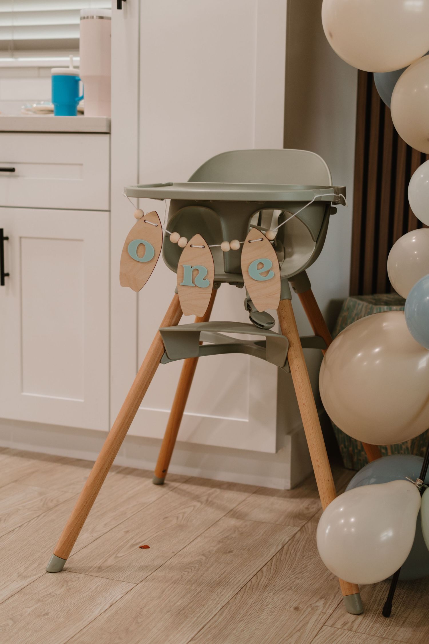 Sage green baby high chair with ONE banner surrounded by blue and white balloon cluster.