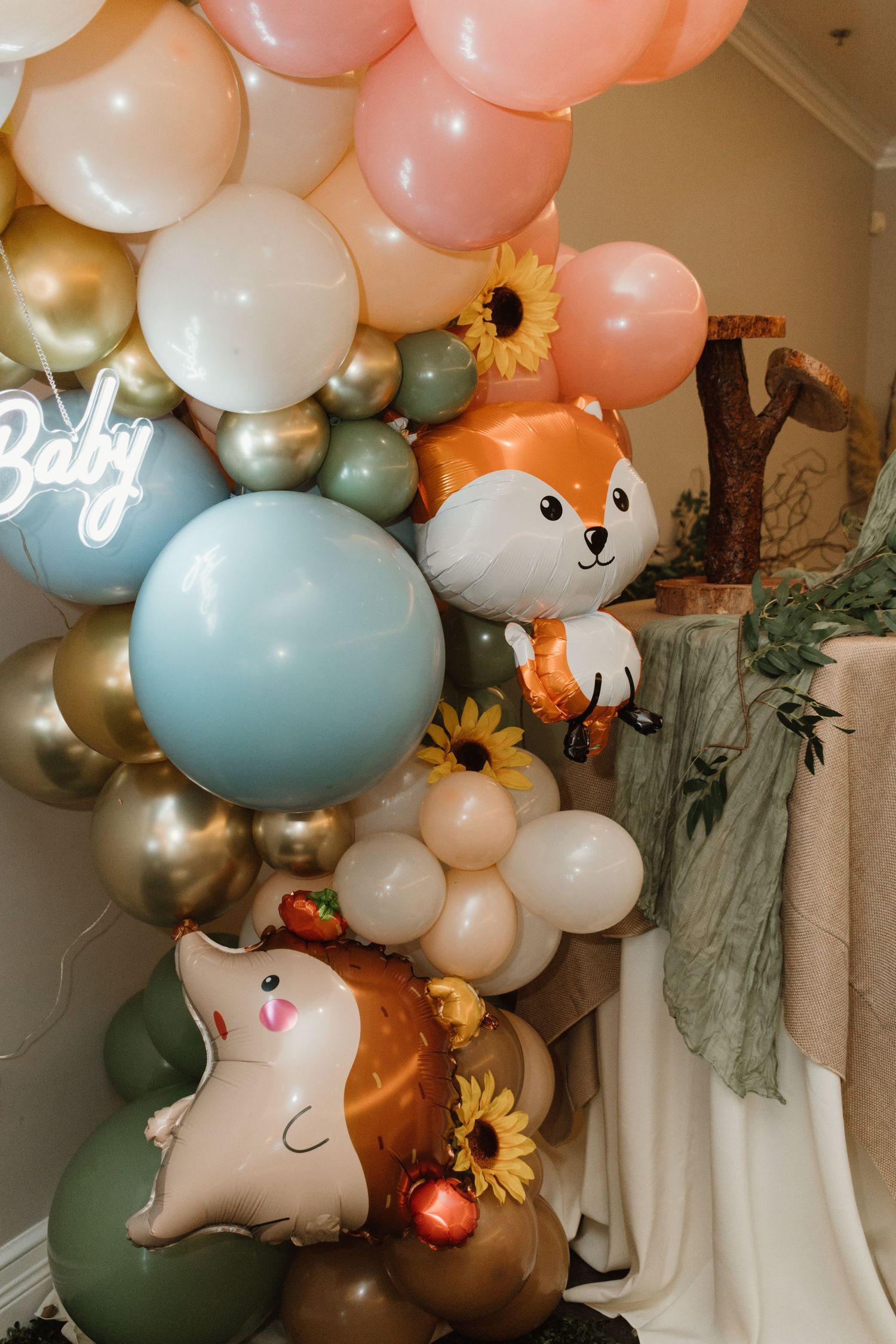 Colorful baby shower balloon garland featuring fox and squirrel foil balloons, sunflowers, and a Baby neon sign.