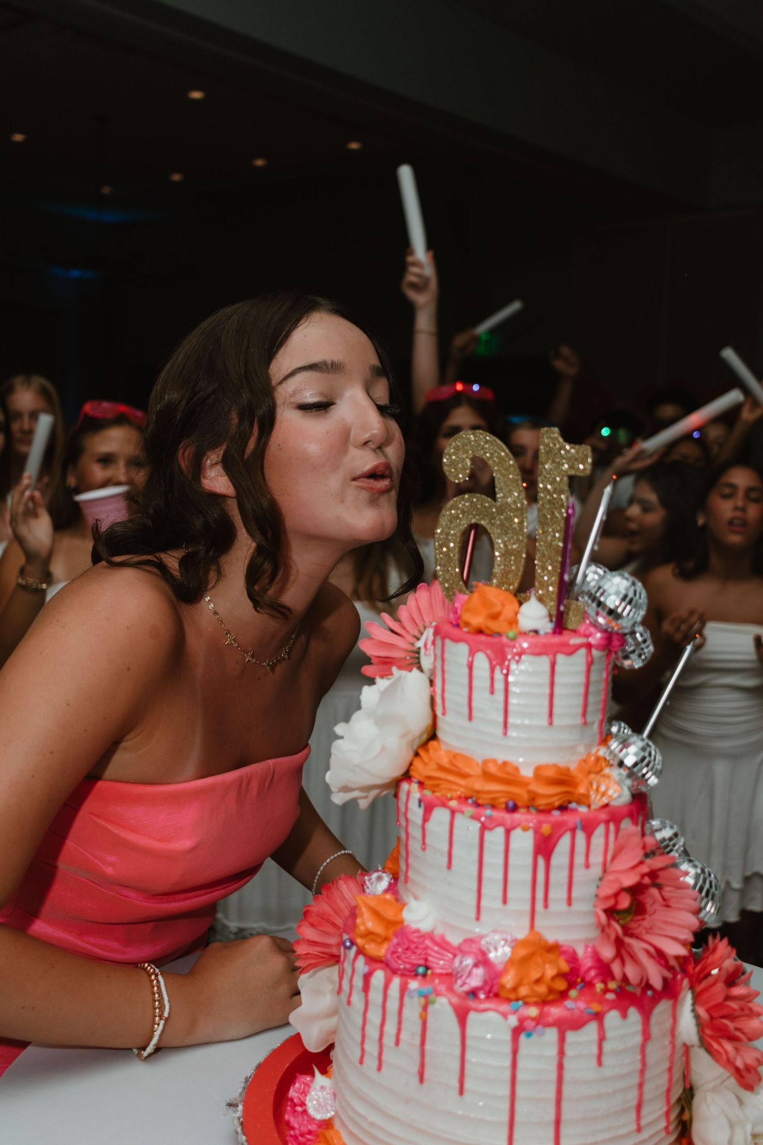 Teen girl blowing out candles on a three-tier pink drip Sweet 16 cake surrounded by celebrating guests.