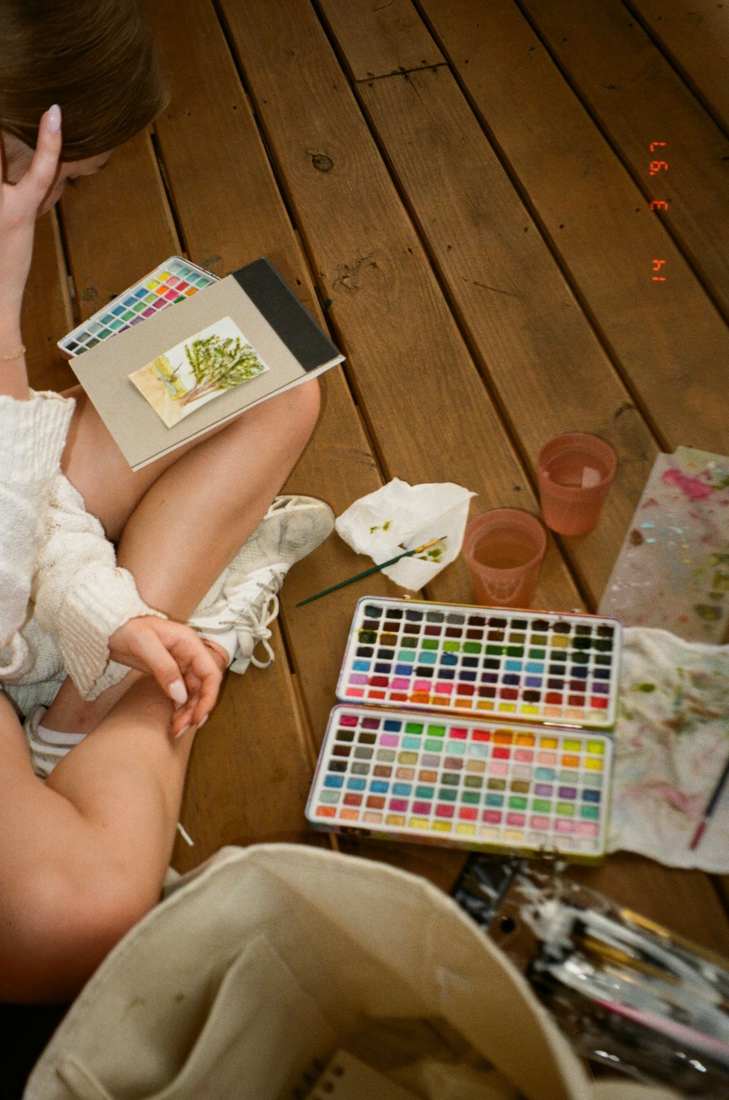 Artist sitting on wooden floor with watercolor palette and art supplies for painting session.