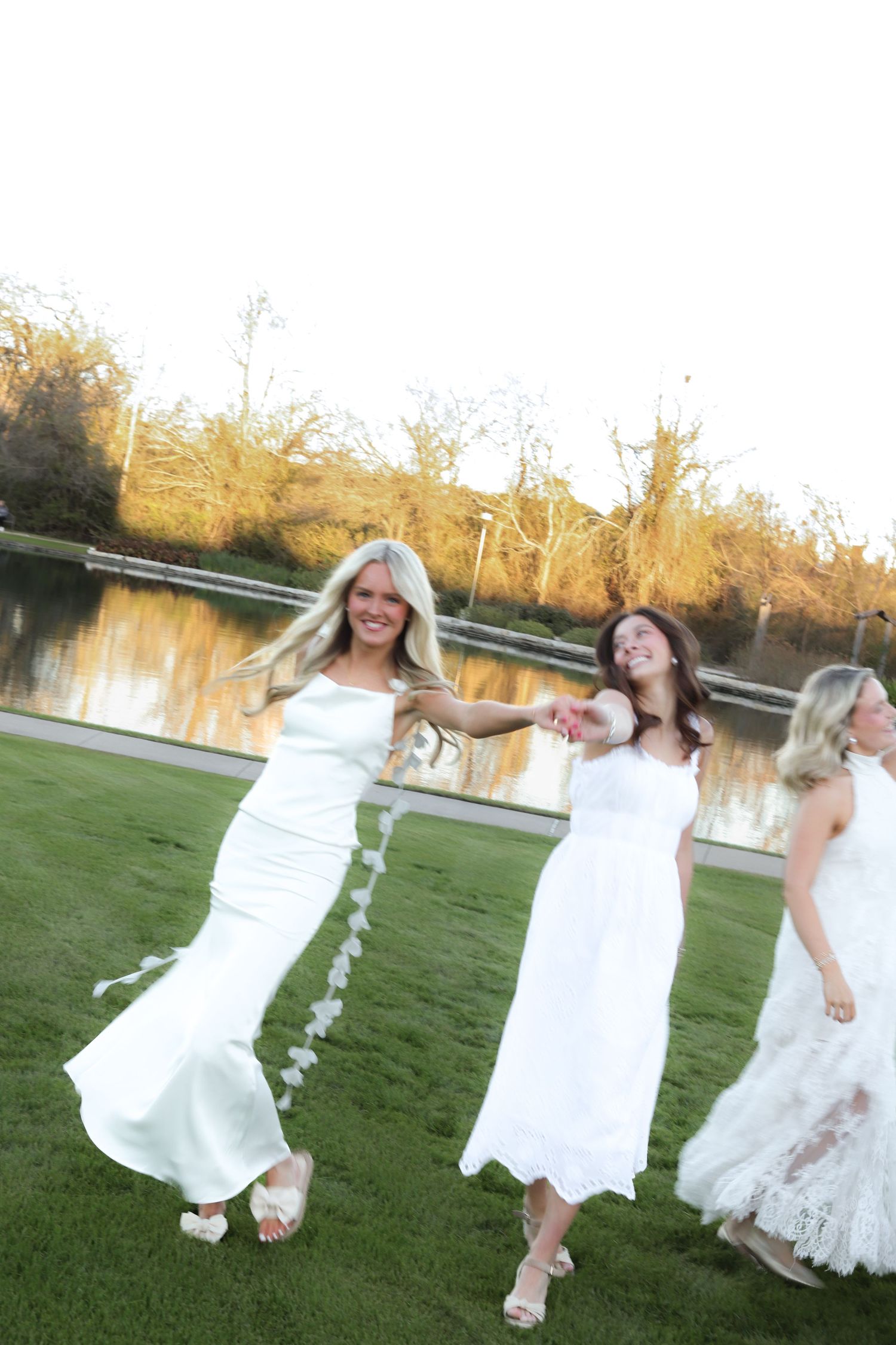 A group of people in white dresses dance and twirl playfully on green grass near a lake at sunset.