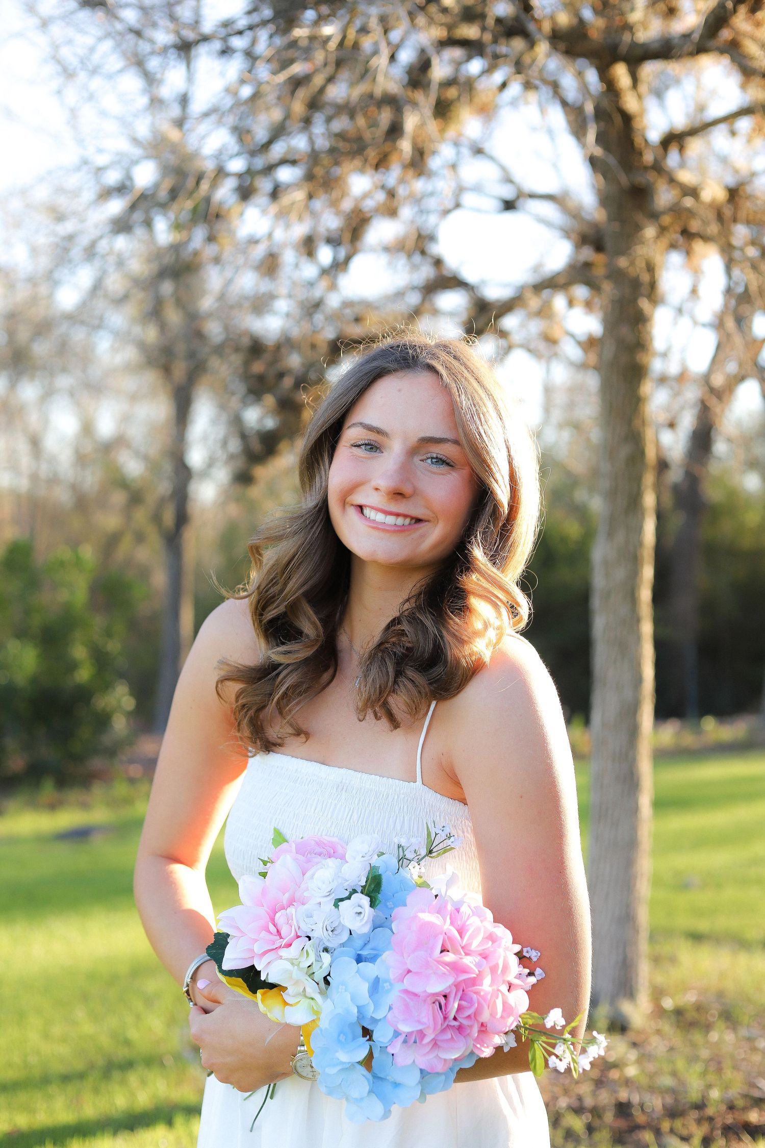 A person in white holds pink and blue flowers while posing in a sunlit outdoor setting.