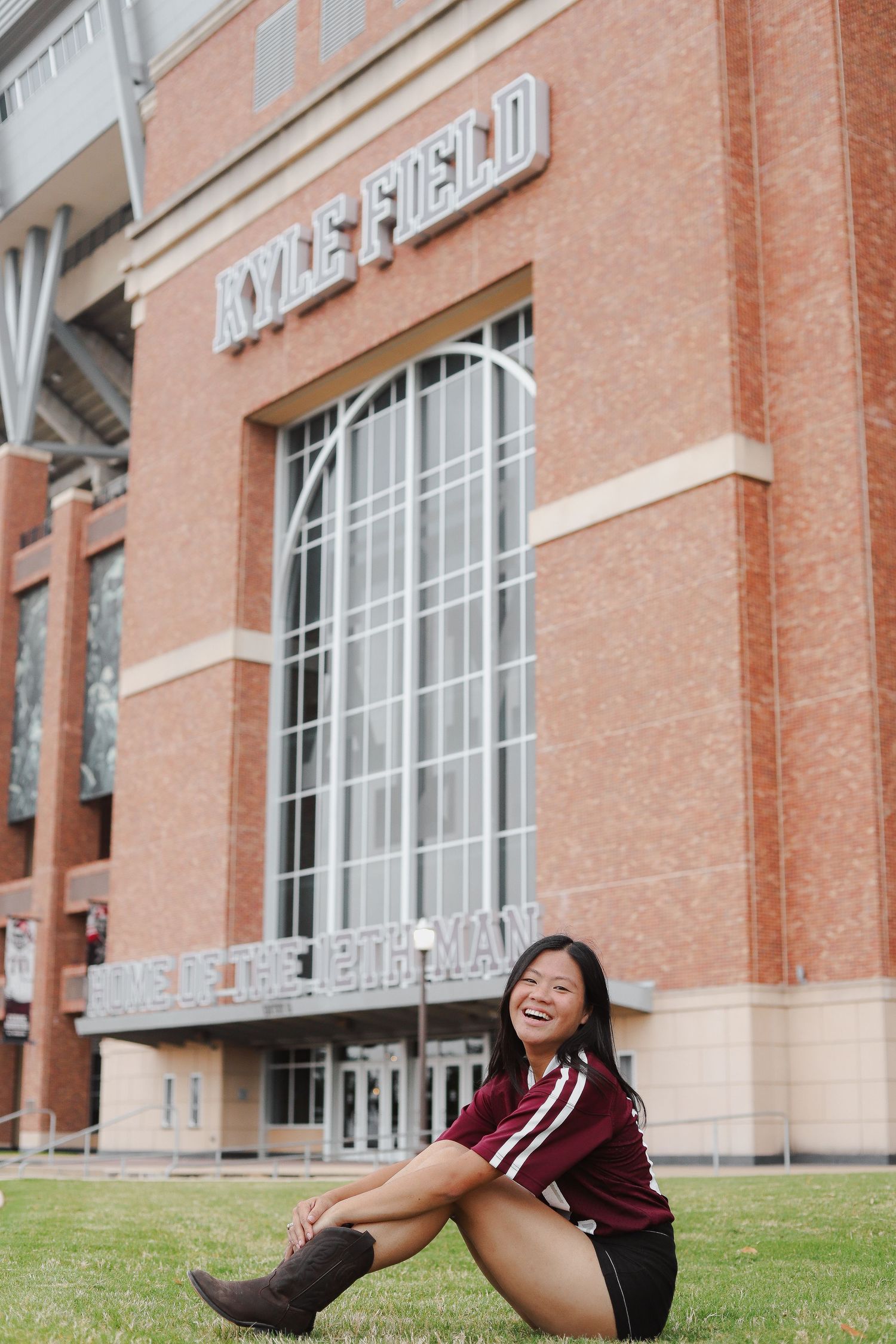 A person in maroon and white sits on grass in front of Kyle Field stadium.