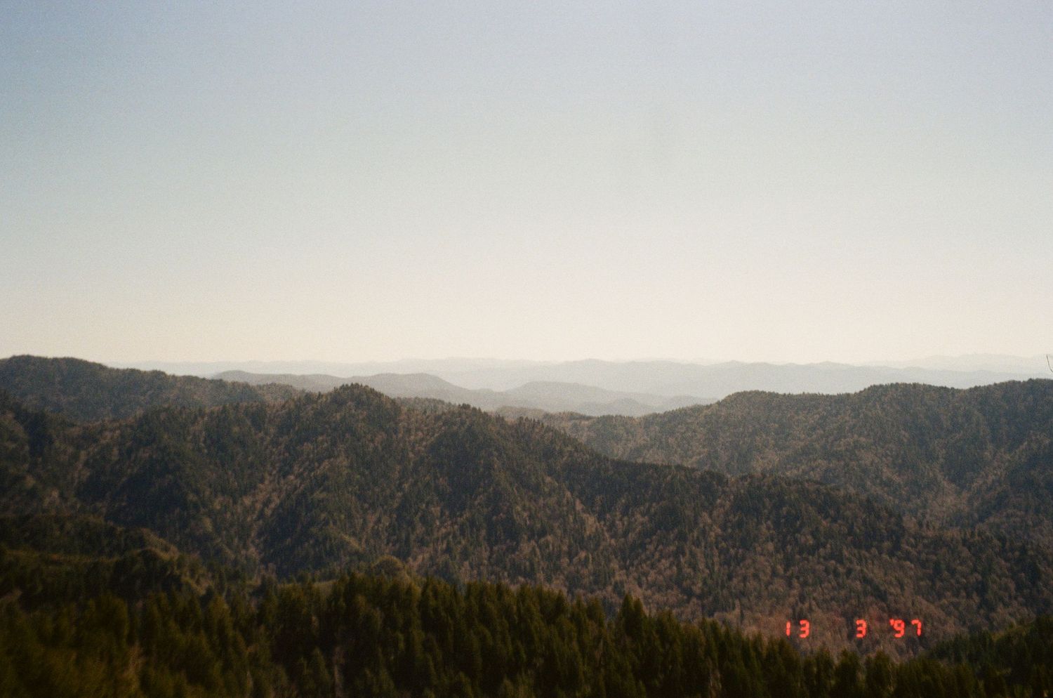A scenic panoramic view of forested mountains and hills stretching across the horizon under an overcast sky.