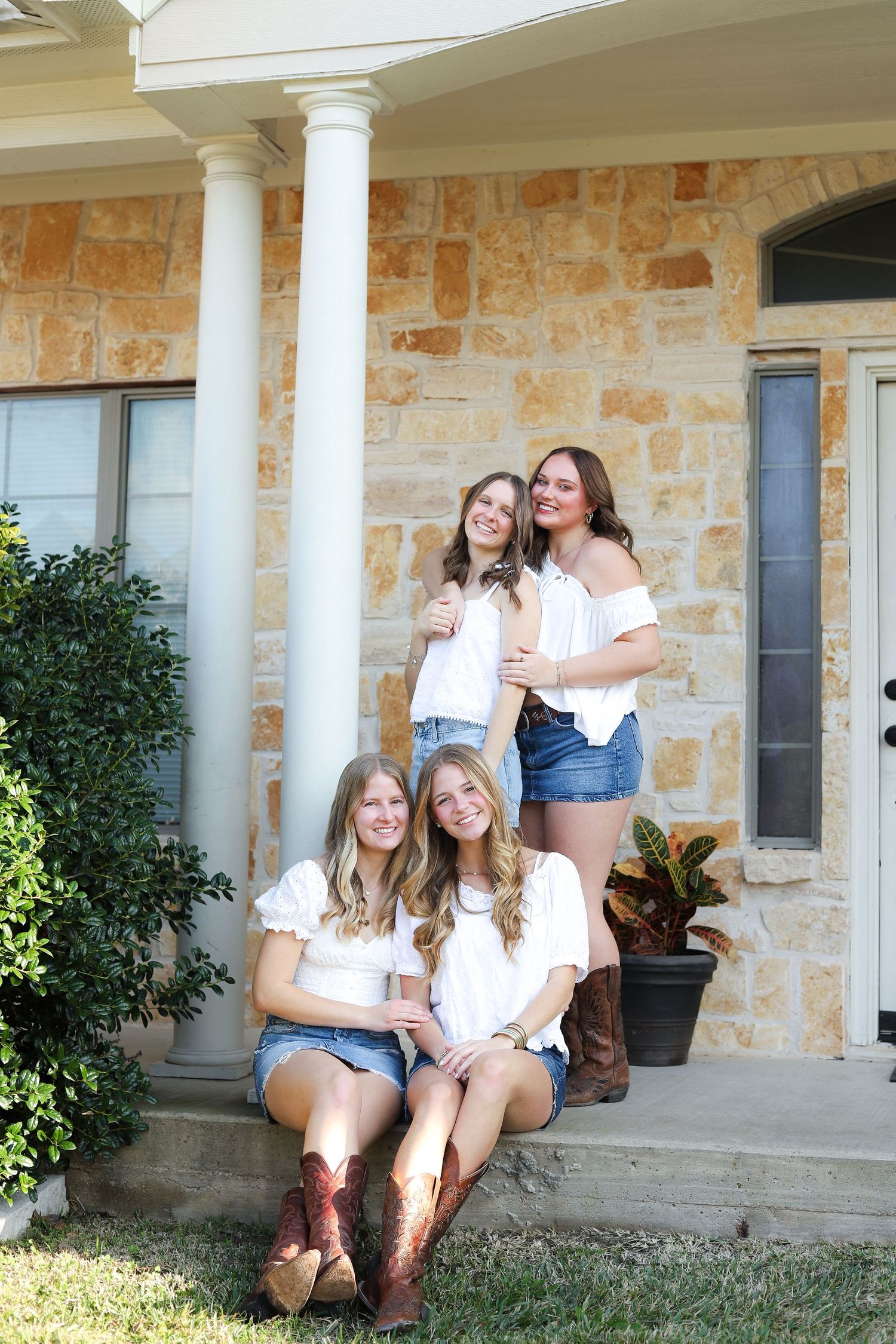 Friends in white tops and denim sit laughing together on a stone porch with columns and potted plants.