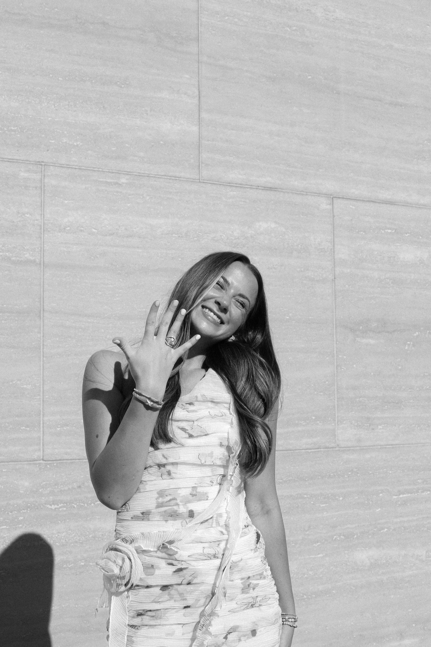 Black and white photo of a woman displaying her ring while smiling against a textured wall.
