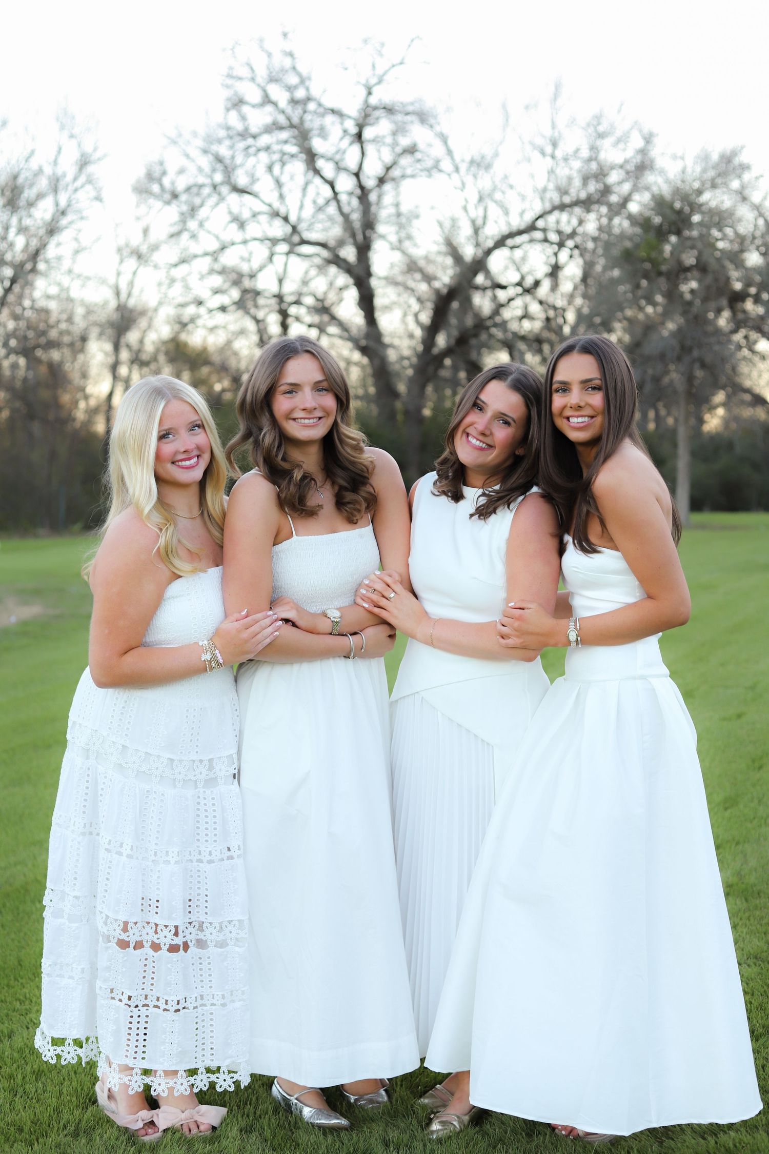 Four women in matching white dresses stand together smiling on a grassy lawn at sunset.