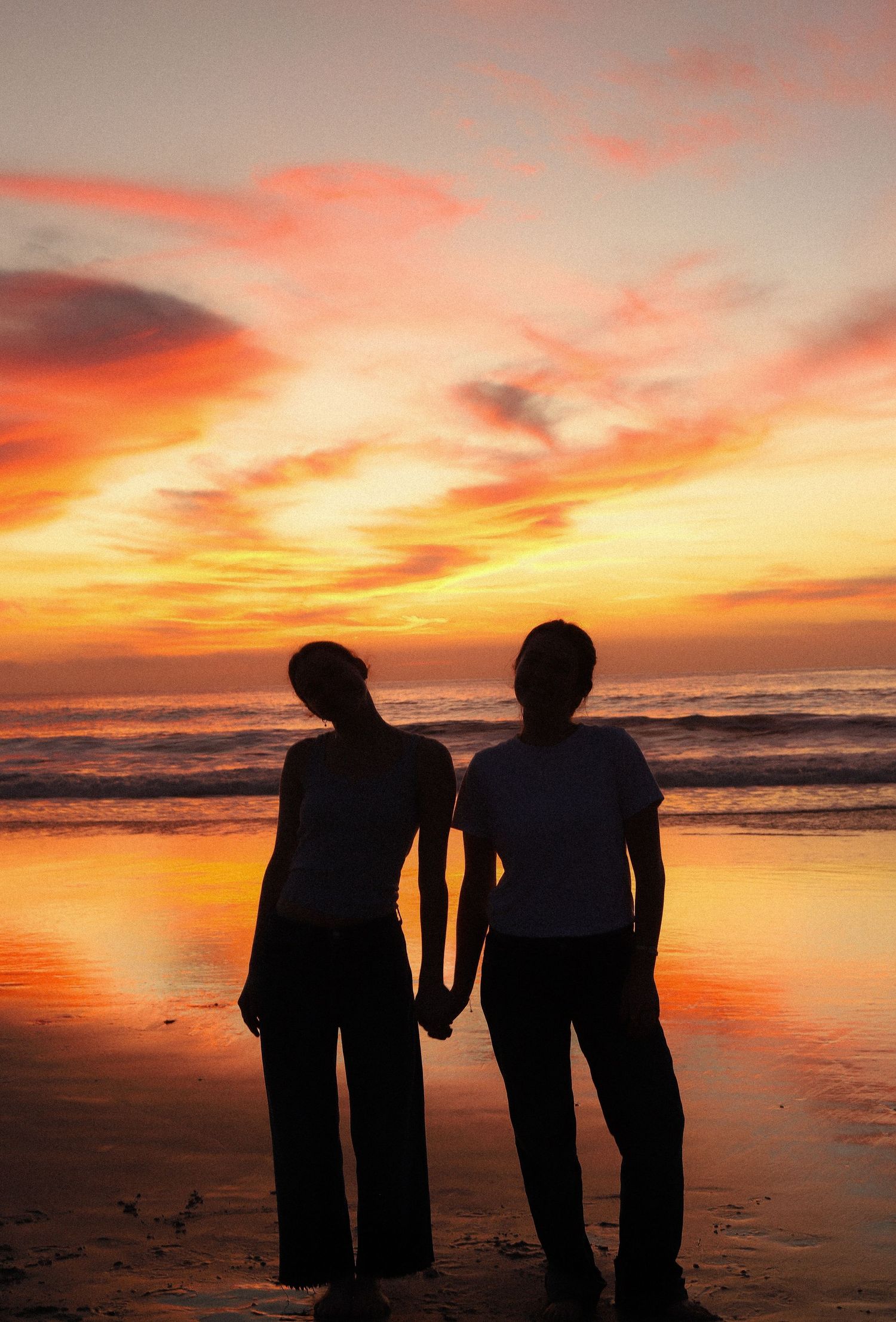 Two silhouettes hold hands while watching a vibrant orange and pink sunset over the ocean from the beach.