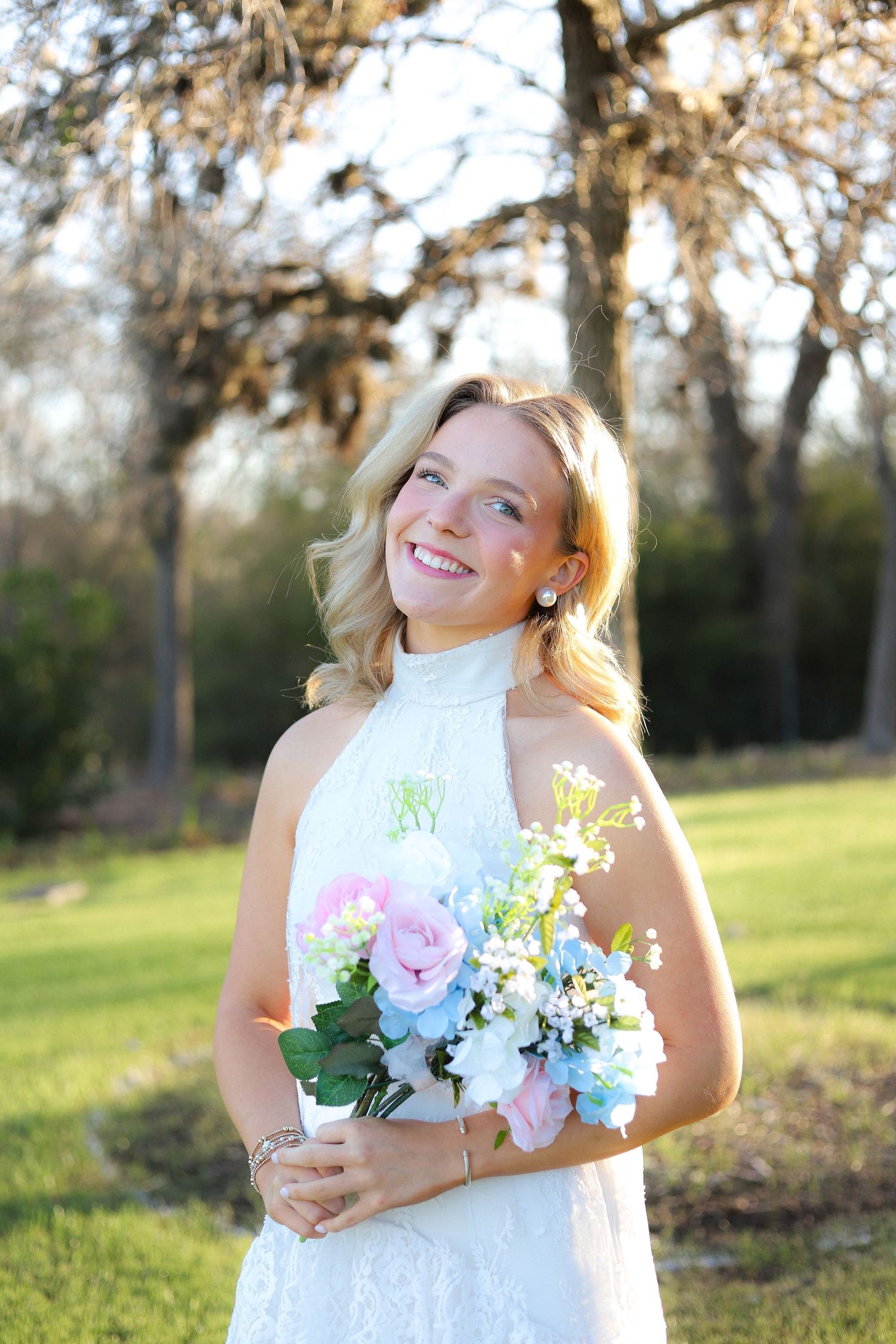 Woman in white lace dress holds bouquet of pink and white flowers in sunlit garden.