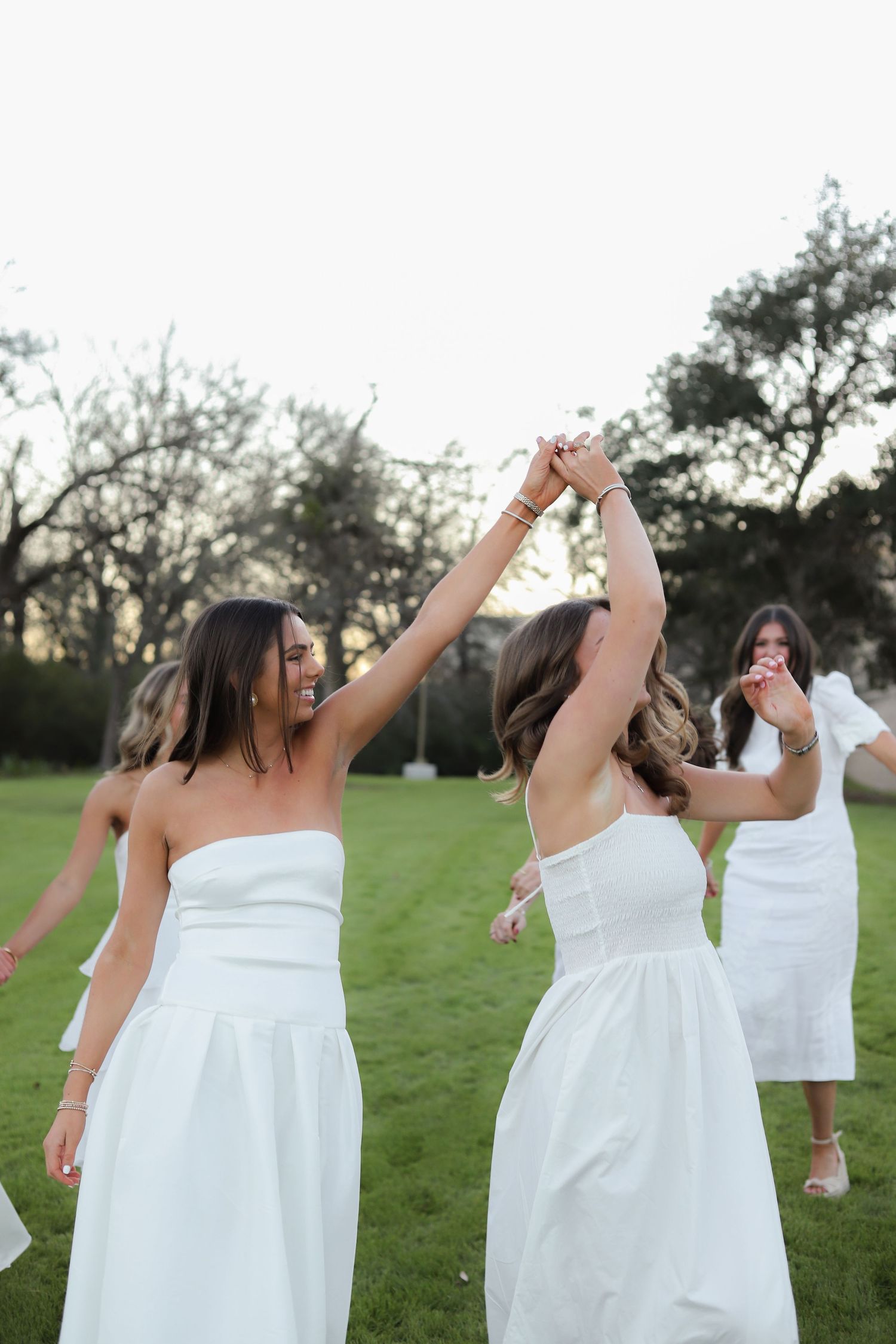 Two people in white dresses dance and twirl together on a lawn at dusk.