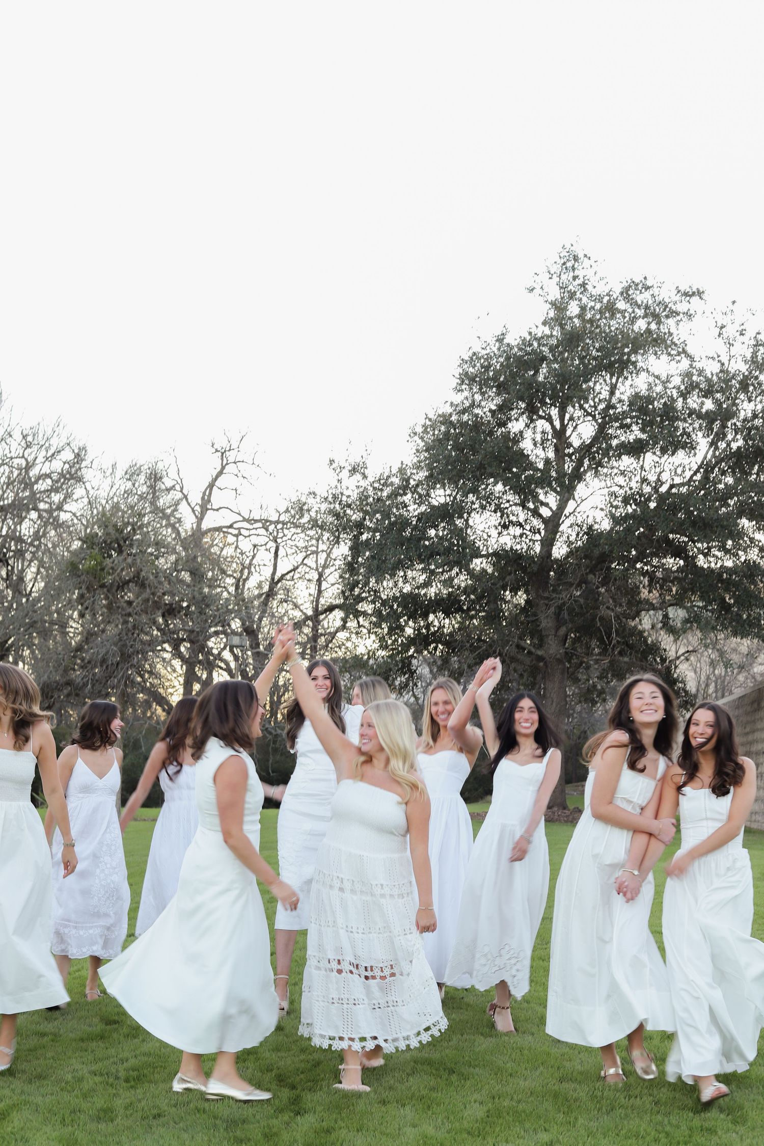 A large group in white dresses poses playfully on grass during a sunset photoshoot.