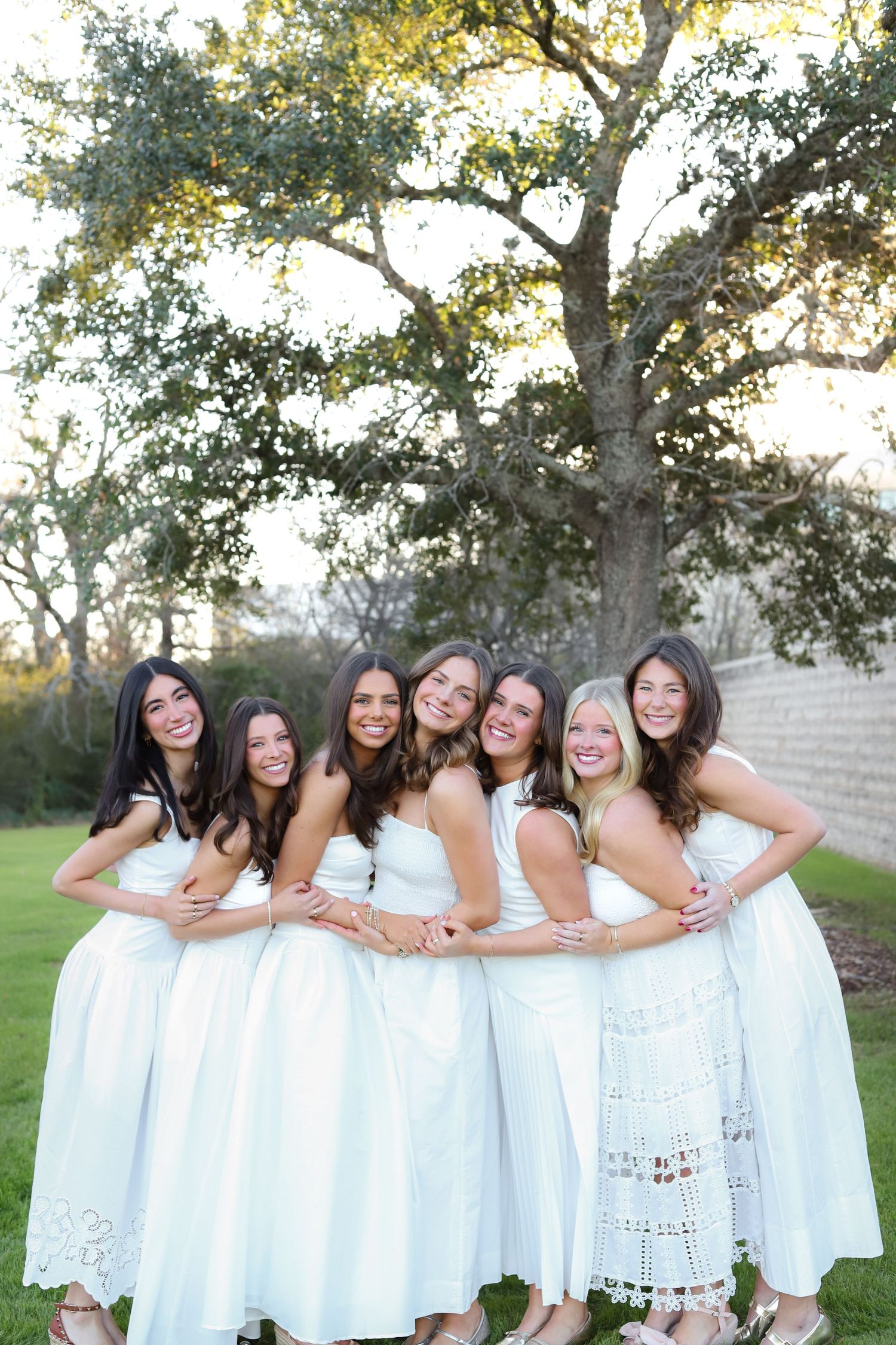 A group in matching white lace dresses pose together outdoors on a sunny day with trees in the background.