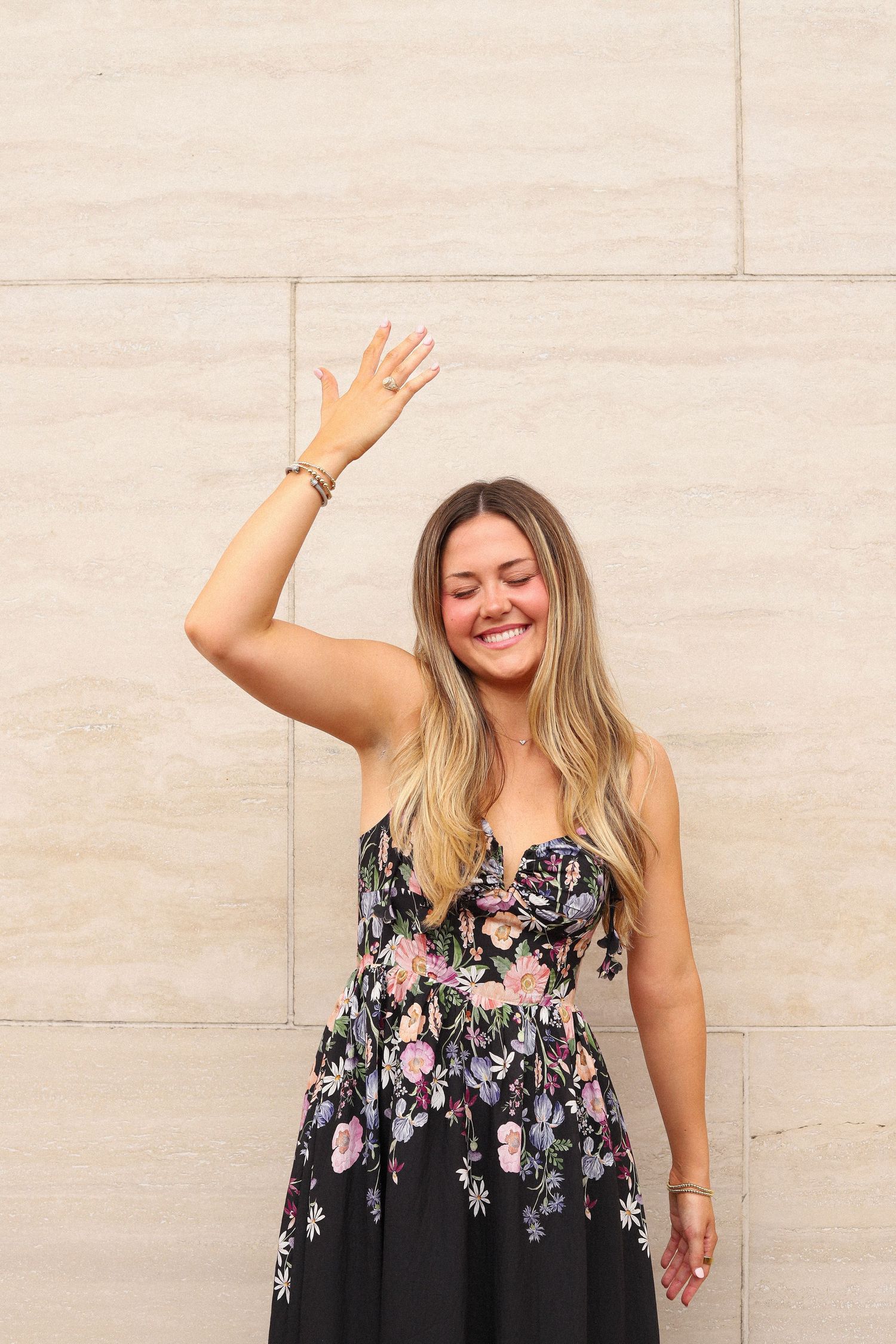 A person in a floral dress raises their hand in greeting against a stone wall.