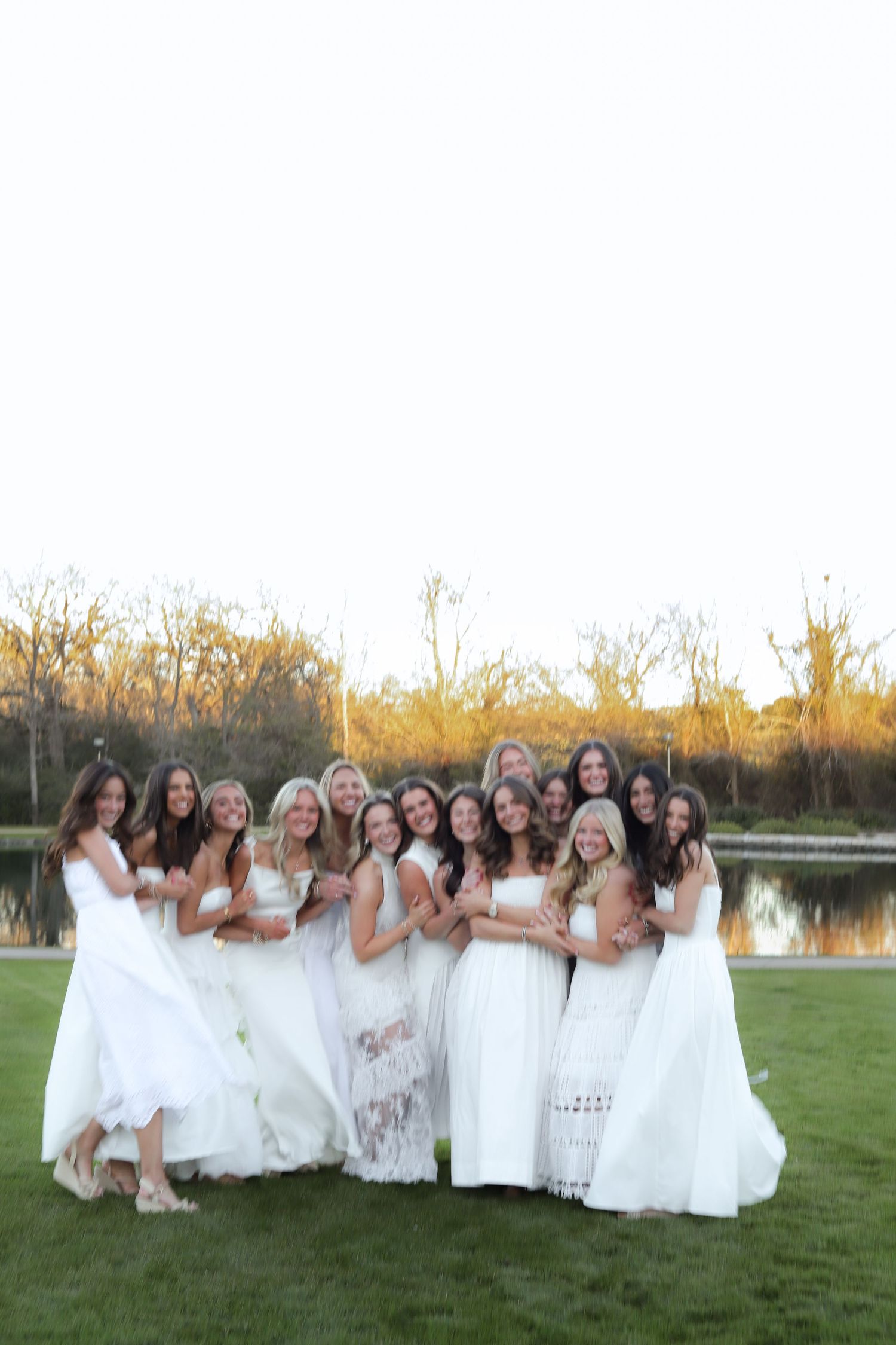 A group in matching white dresses poses together on grass near a lake at sunset.