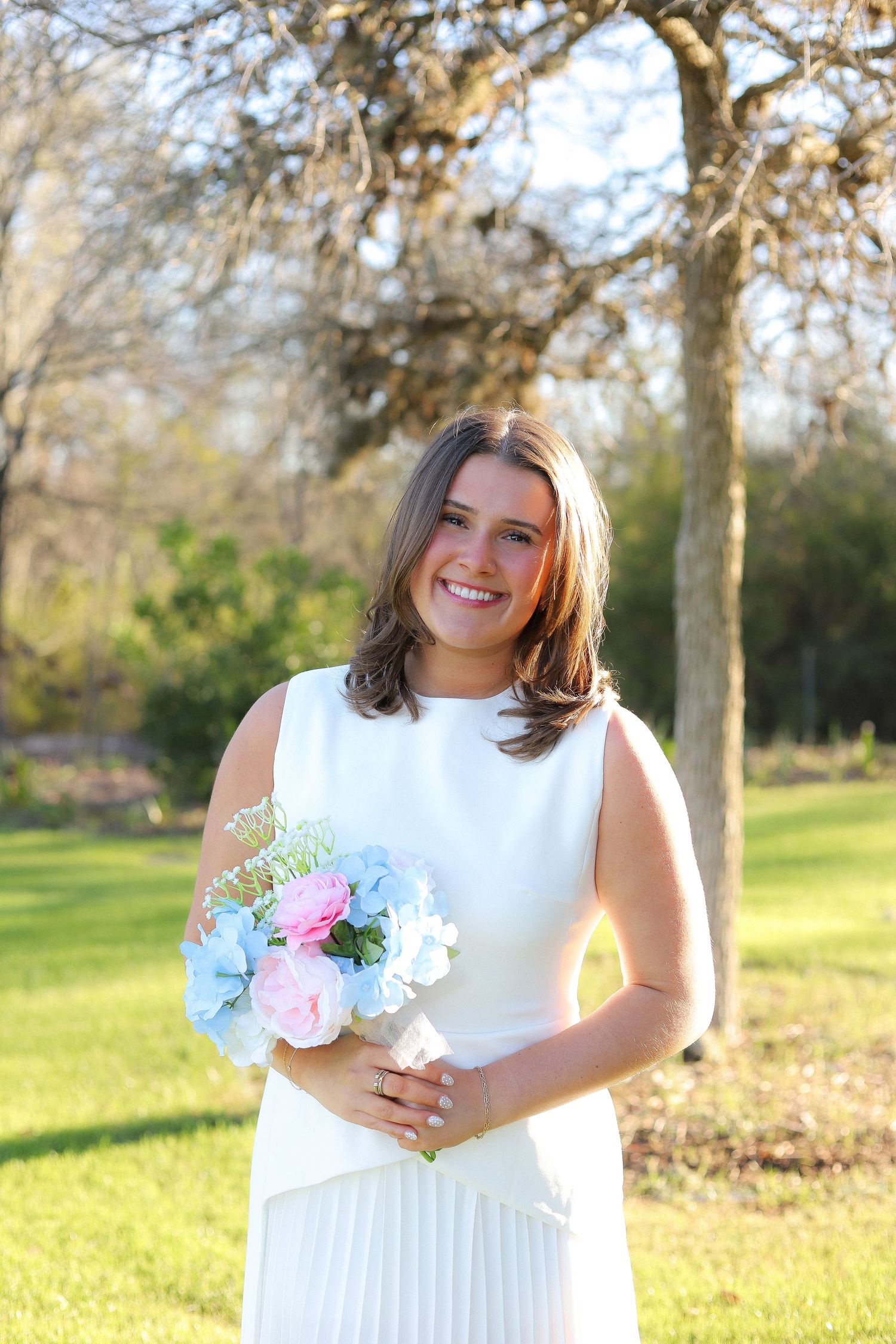 A person in a white dress holds a pastel floral bouquet while standing in a sunny park setting.