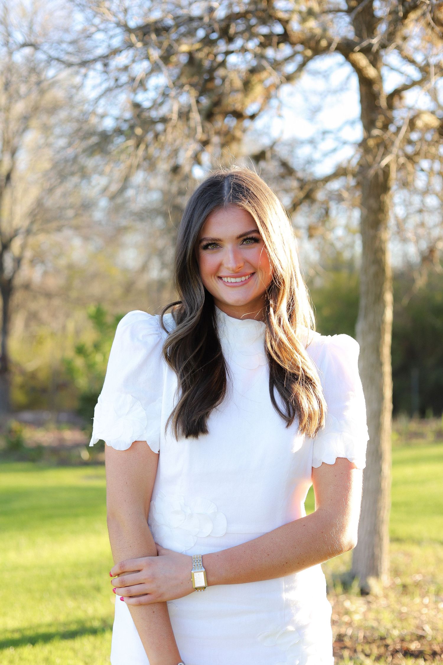 A person in a white puff-sleeve dress poses outdoors in a sunlit park setting.