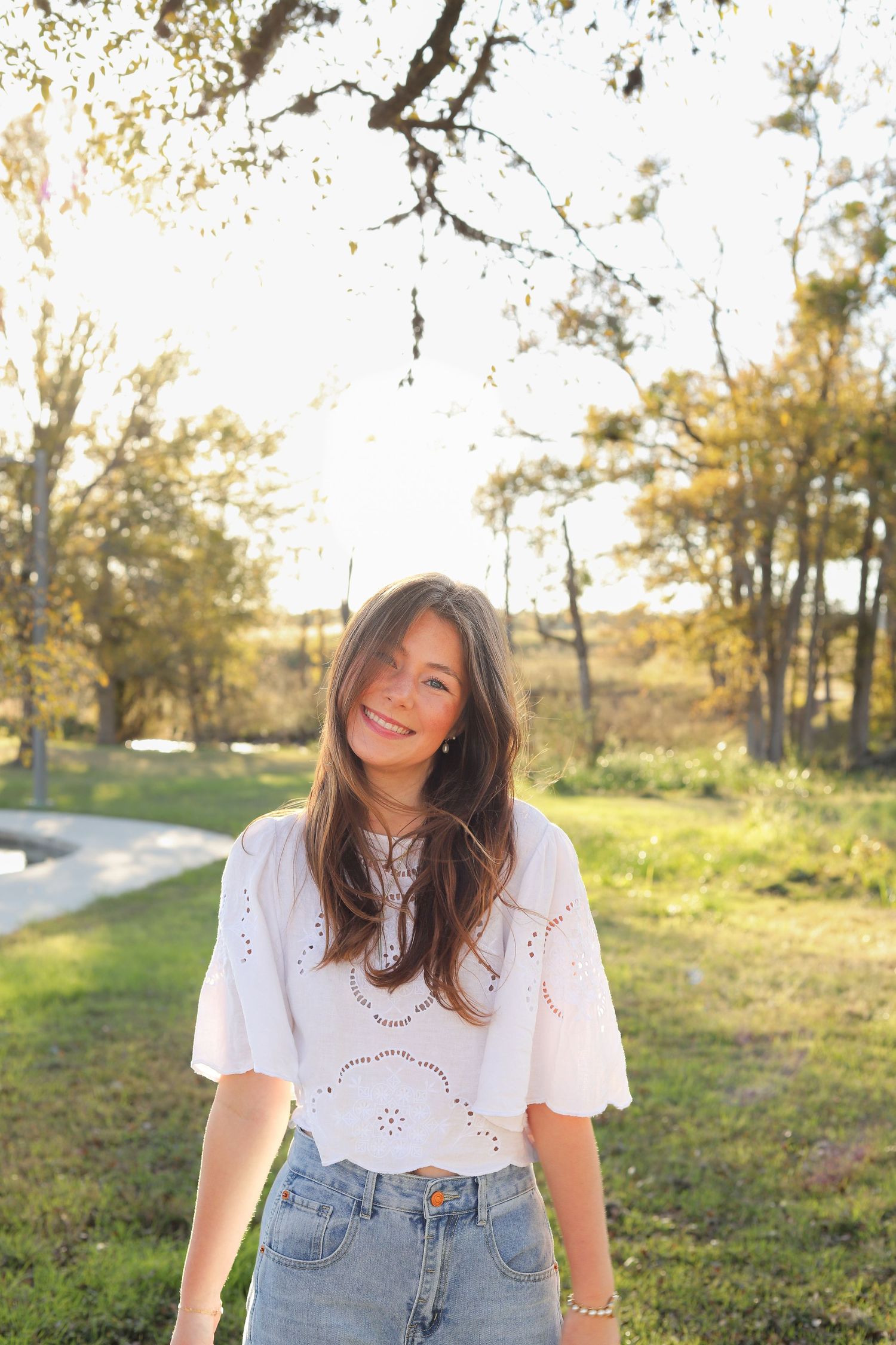 A joyful portrait in a white eyelet top and jeans against a sunlit natural backdrop with trees.