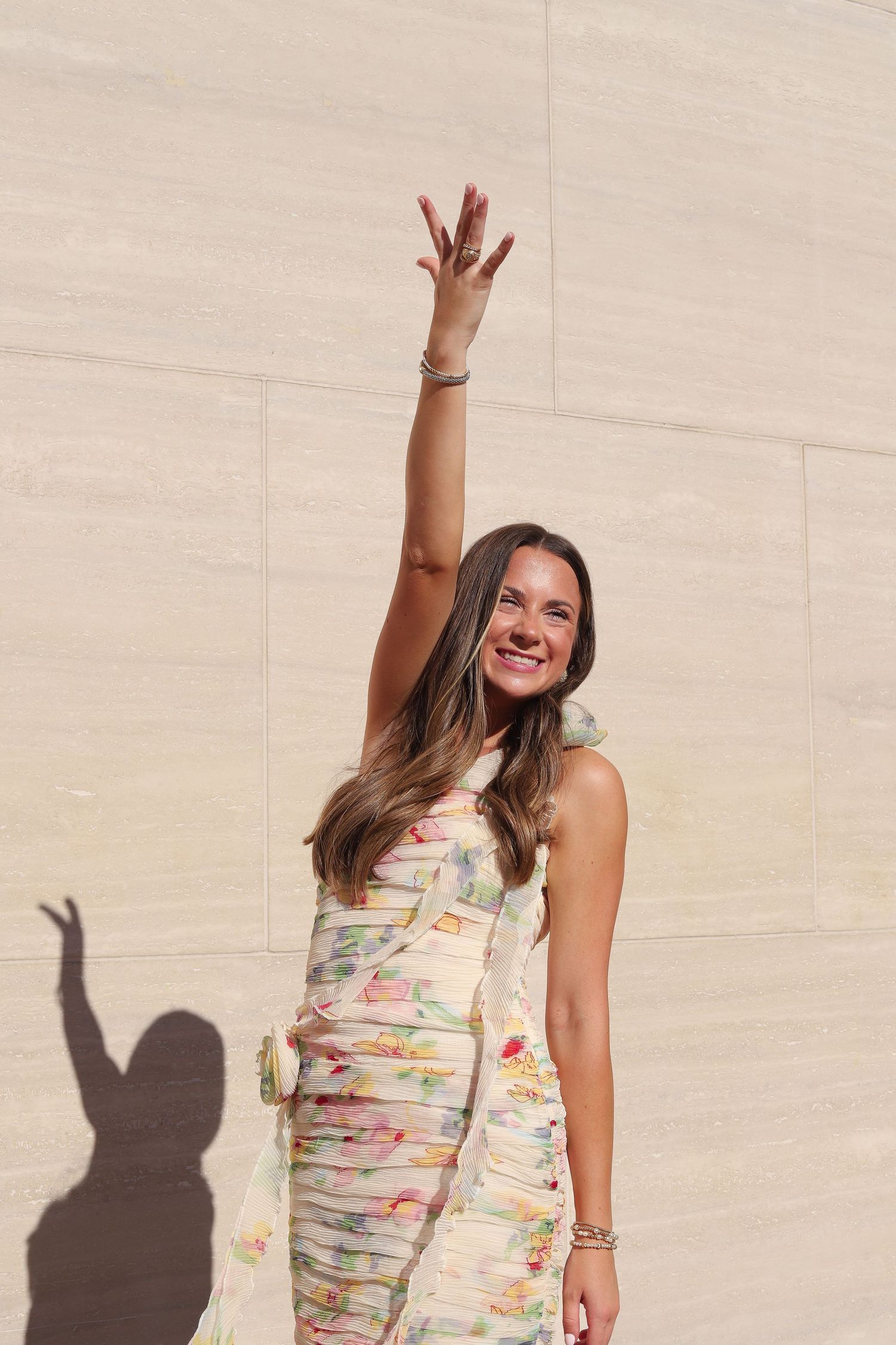 A woman in a fitted dress raises her arm joyfully against a stone wall with dramatic shadows.