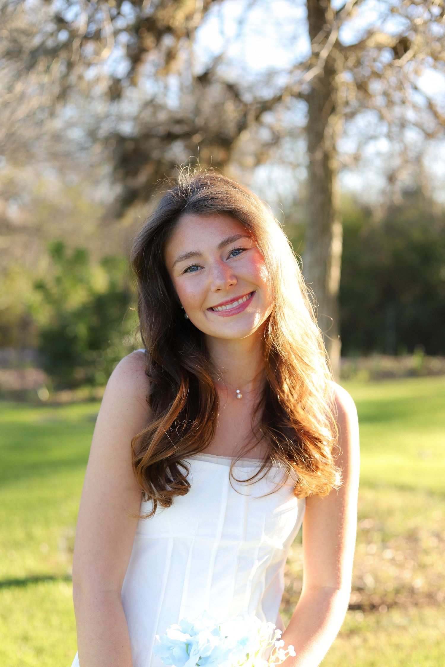 A person in a white sleeveless top smiles joyfully while posing outdoors in golden sunlight.