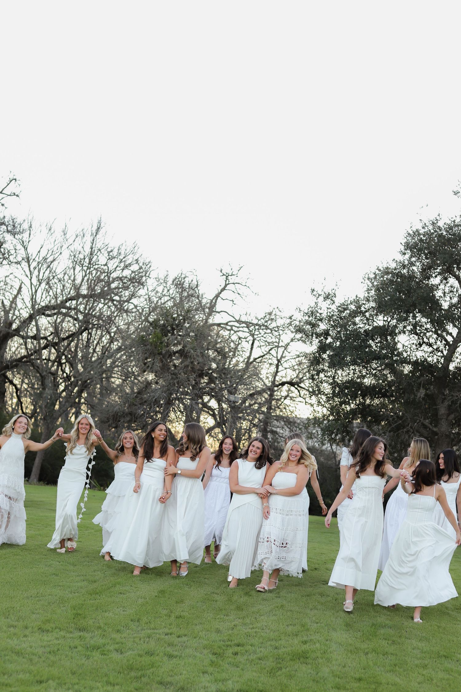 A group of women in flowing white dresses dance together on a grassy lawn surrounded by trees at dusk.