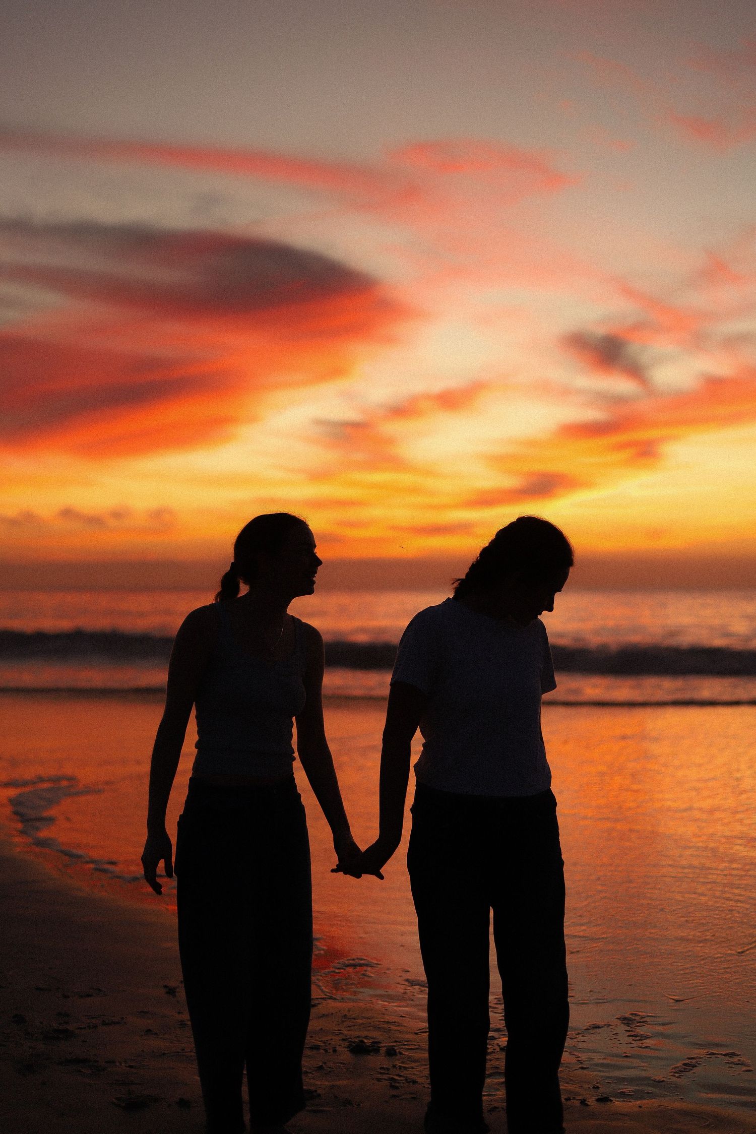 Two silhouettes hold hands on a beach at sunset with vibrant orange and red clouds reflecting on the ocean.