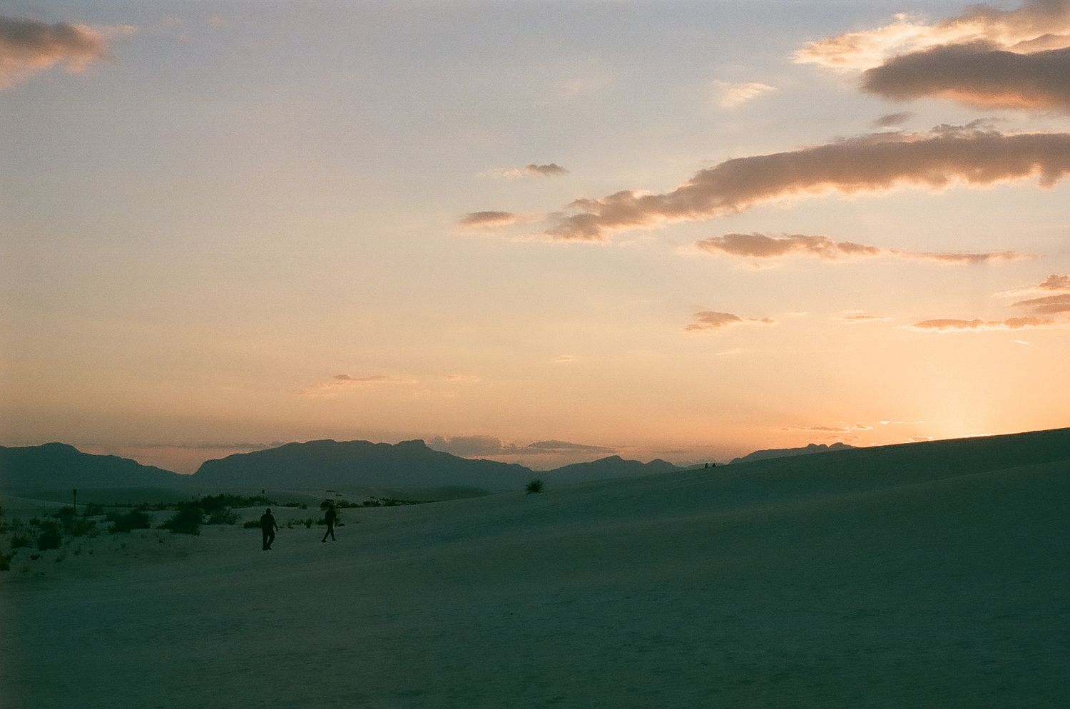 Silhouettes of people walking across sand dunes at sunset with mountains in the distance and orange clouds above. White sands New Mexico