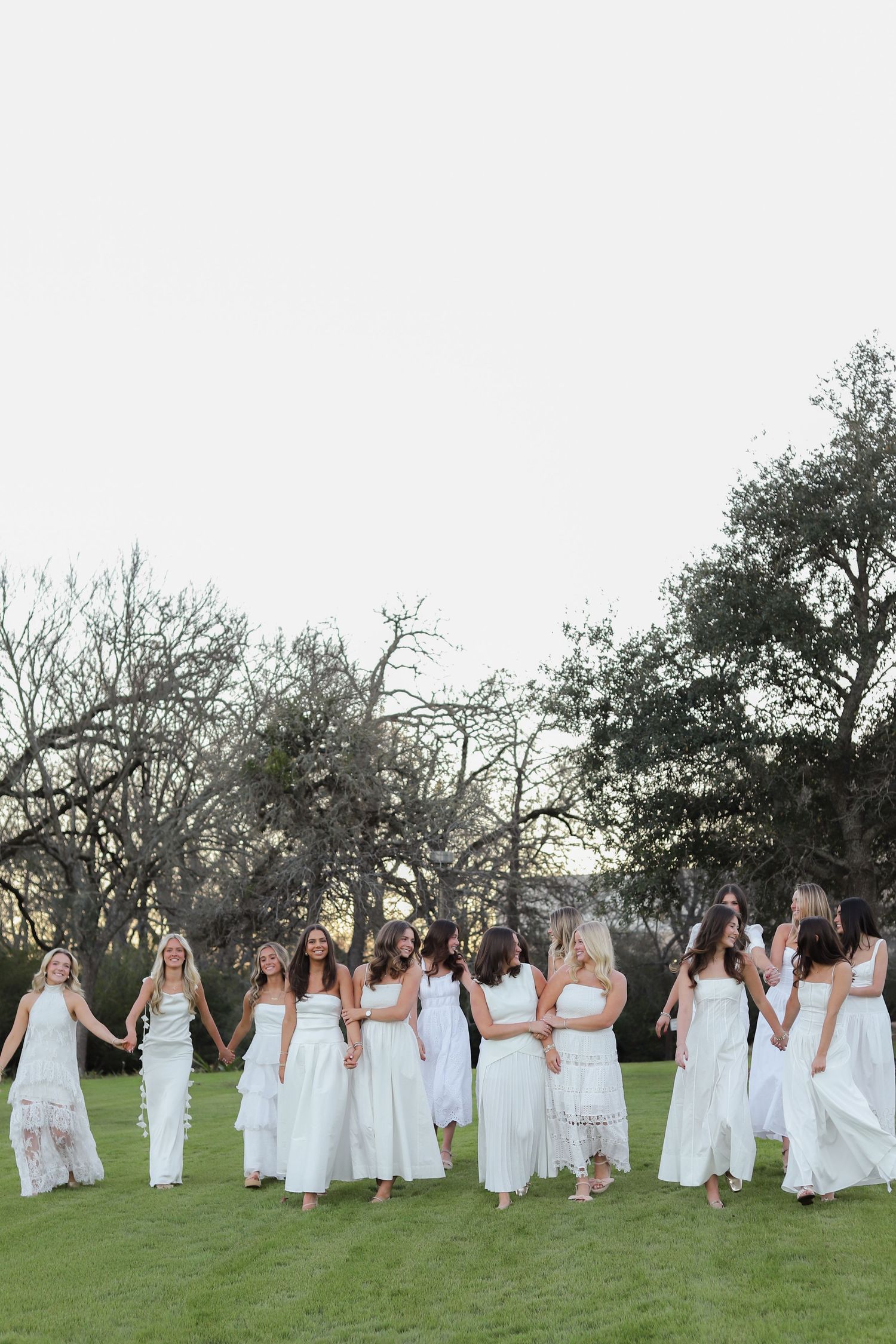 A line of people wearing white dresses hold hands while standing in a grassy field at dusk.