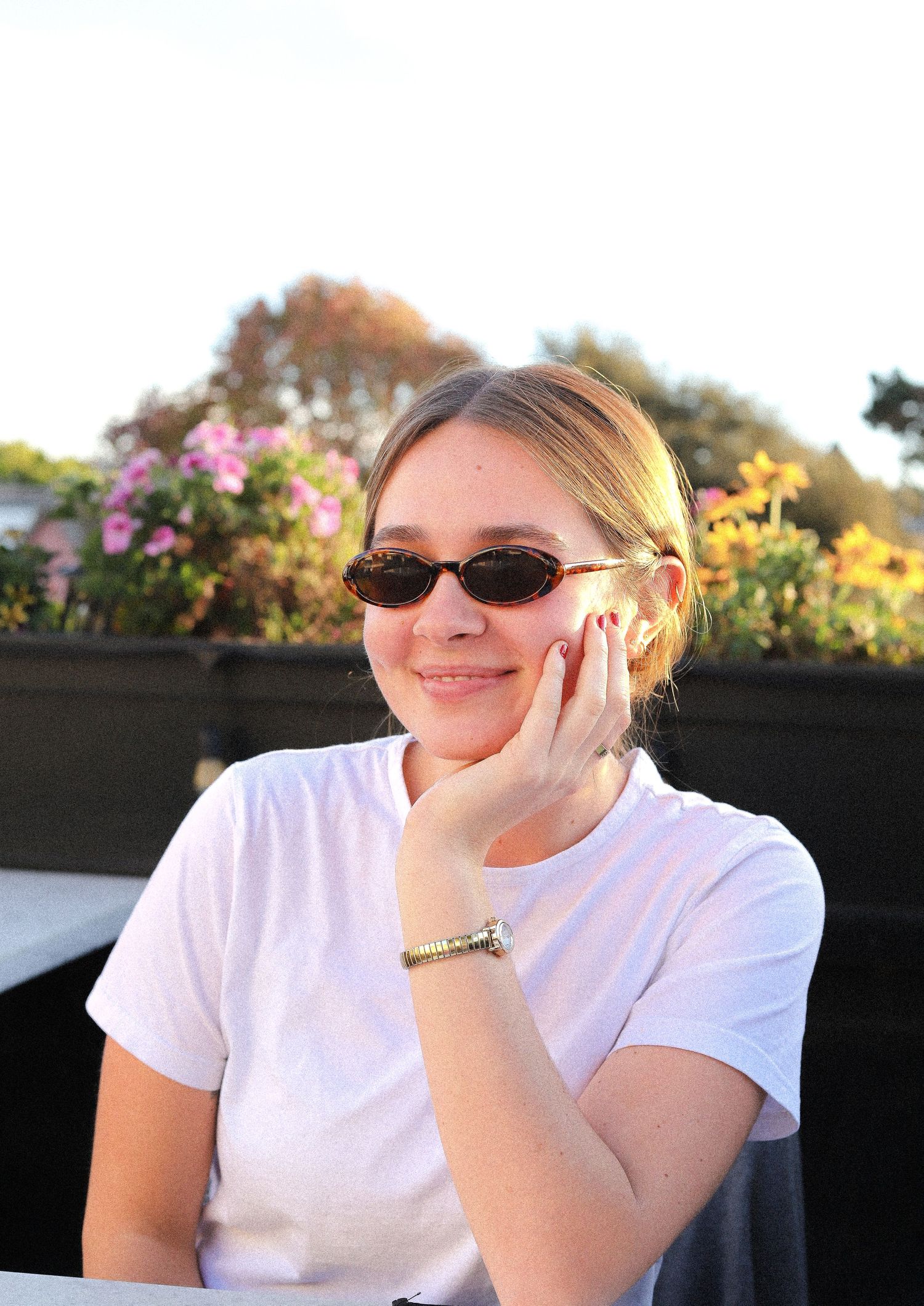 A person wearing sunglasses and a white t-shirt sits outdoors with colorful flowers blooming in the background.
