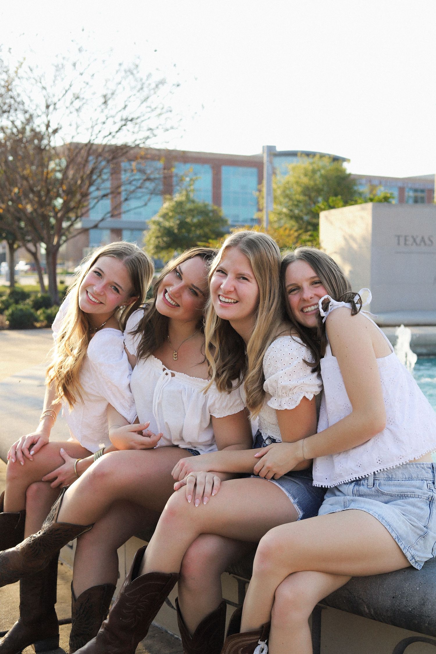 Four friends in white tops pose together on a ledge at Texas A&M University with a building in the background.