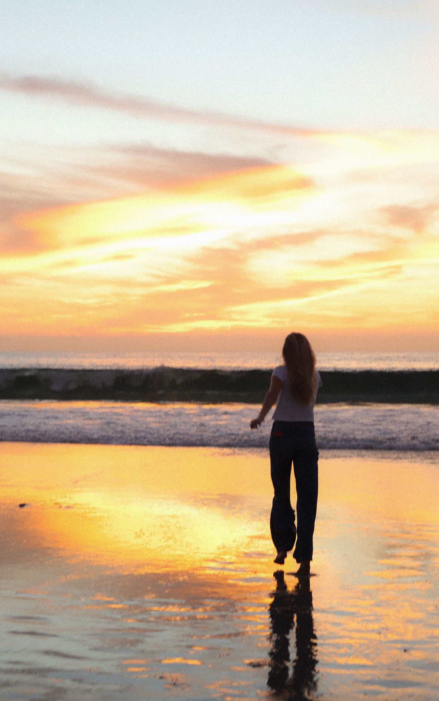 A silhouette stands on a reflective beach at sunset with golden clouds and waves in the background.